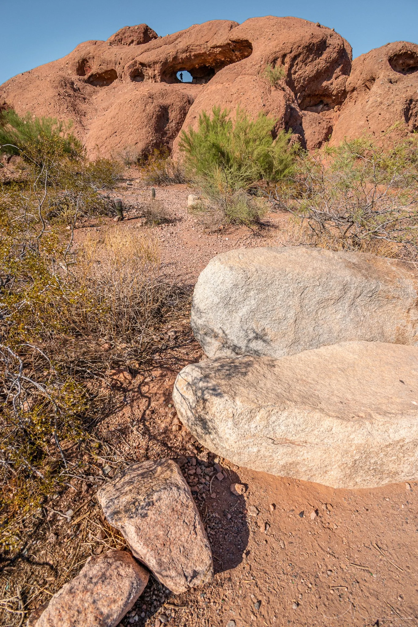 Arid desert landscape with large reddish-brown rock formations, sparse bushes, and a clear blue sky.