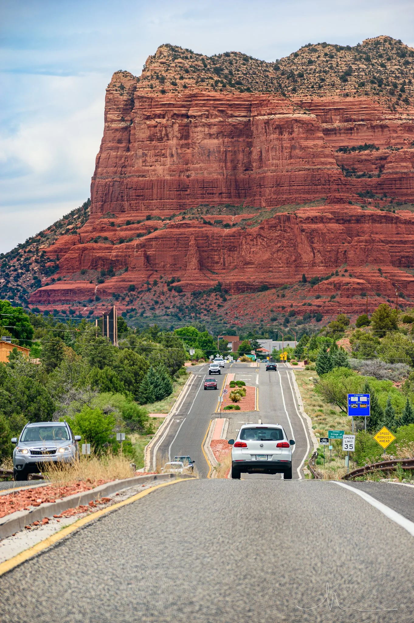 A scenic view of a street flowing towards a red rock formation with mountains, with parked and moving cars, trees, and road signs.