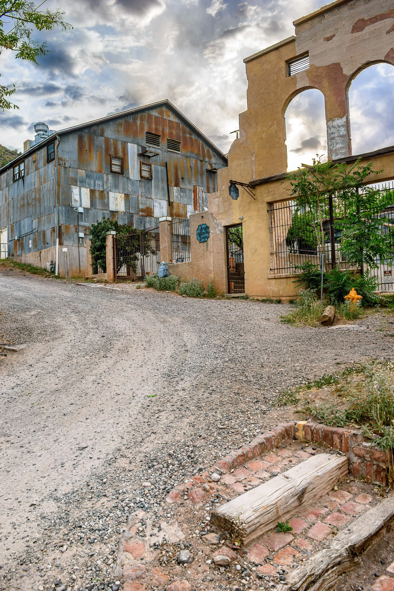 Rustic alleyway with a gravel road leading towards old buildings, one with weathered metal siding and the other with a stucco finish, under a cloudy sky.