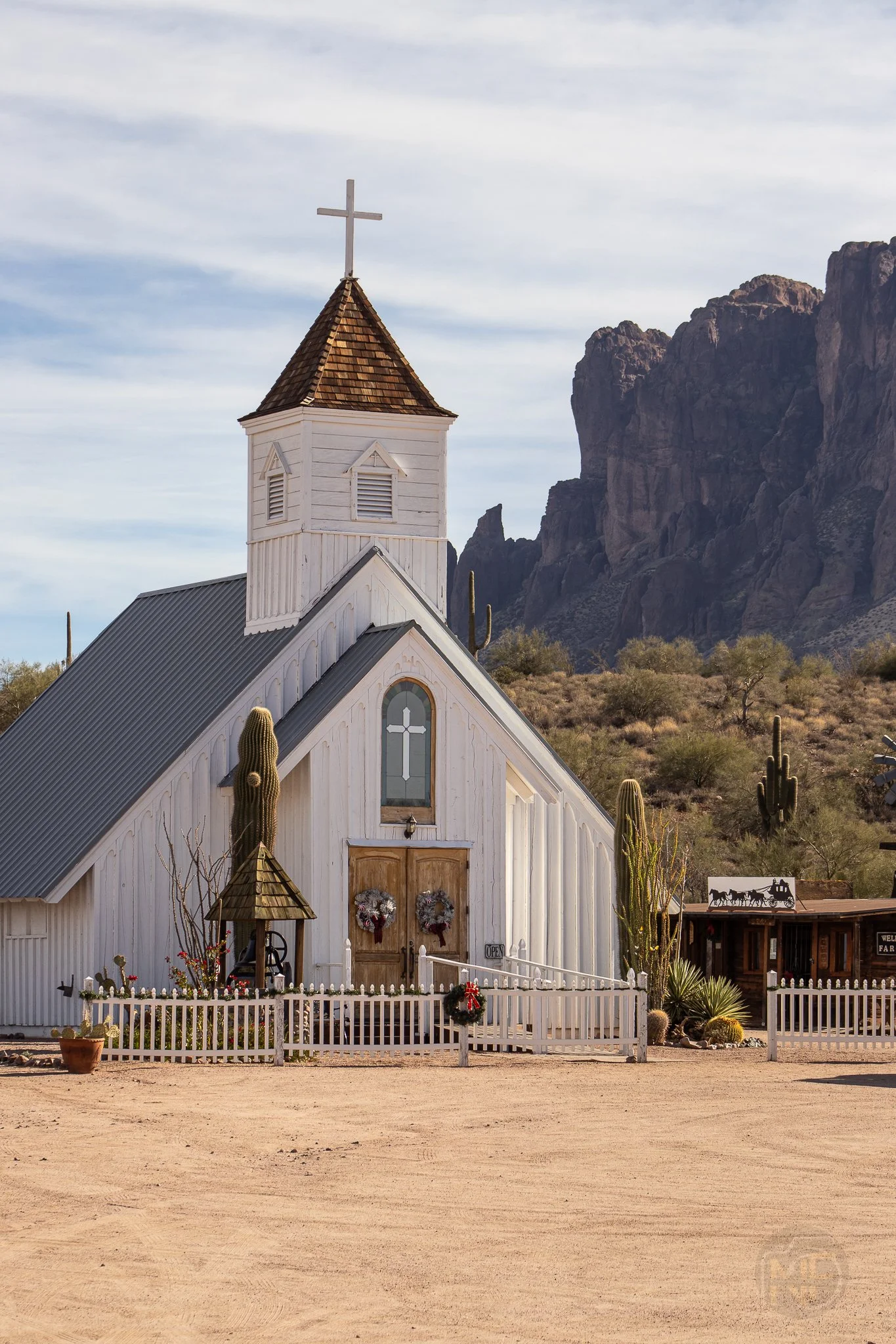 A small white church with a brown-shingled steeple, cross on top, set in a desert landscape with cacti and mountains in the background.