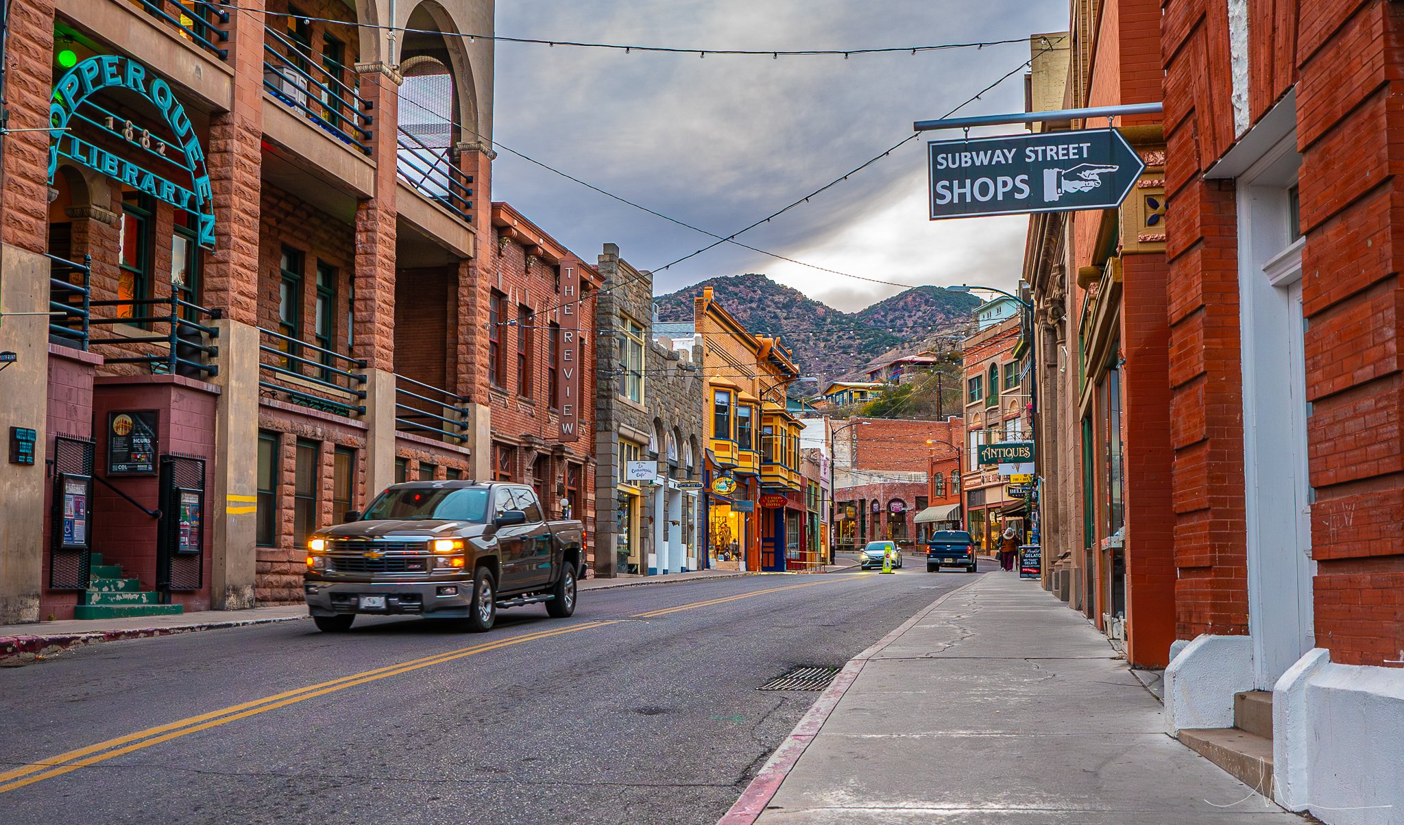 A street scene in a small town with brick buildings, shops, and cars. Mountains visible in the background. Sign for Subway Street Shops and other storefront signs.