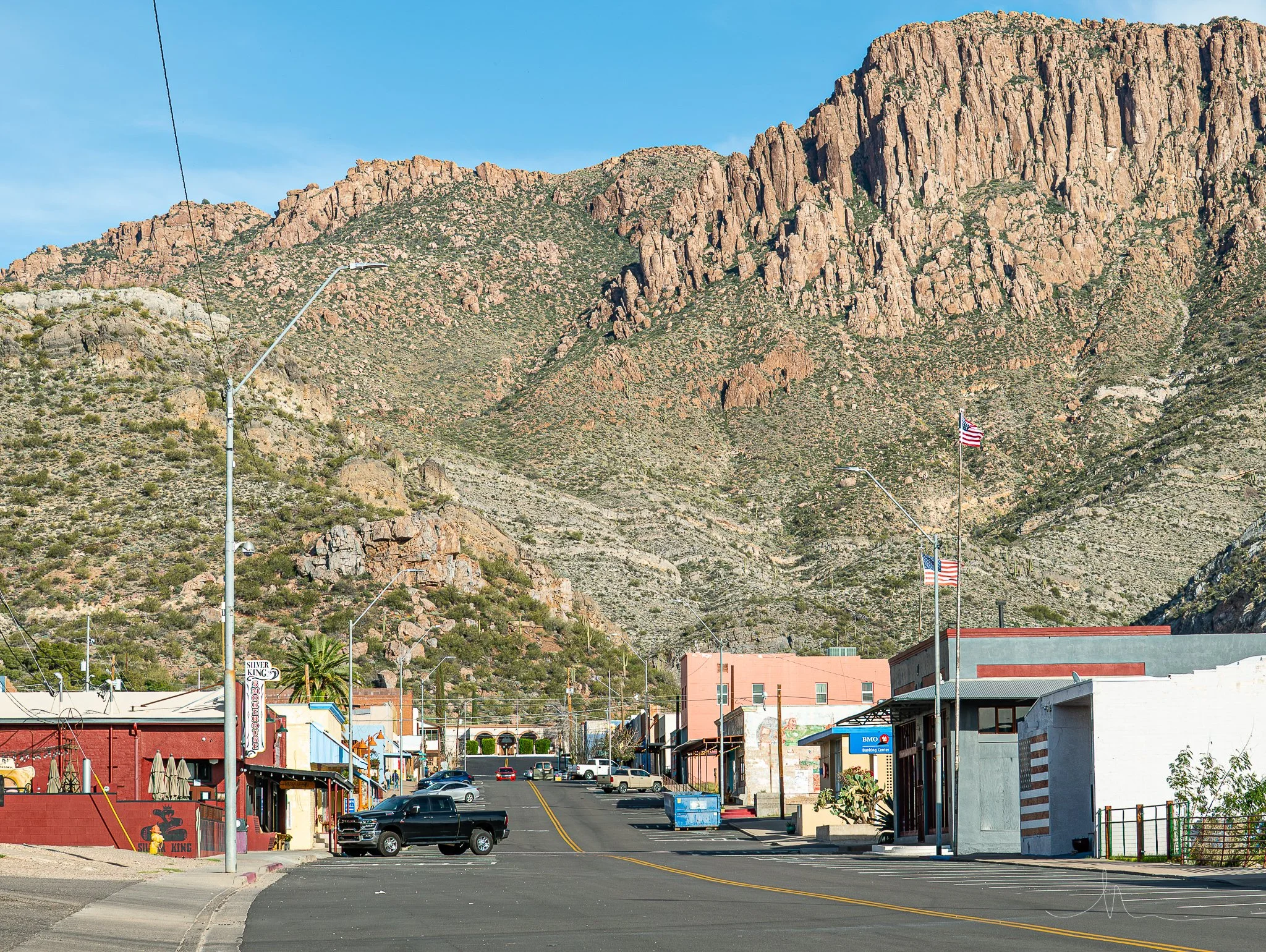 Small town street with colorful buildings and American flags, set against a mountainous landscape with rocky cliffs and sparse vegetation under a clear blue sky.