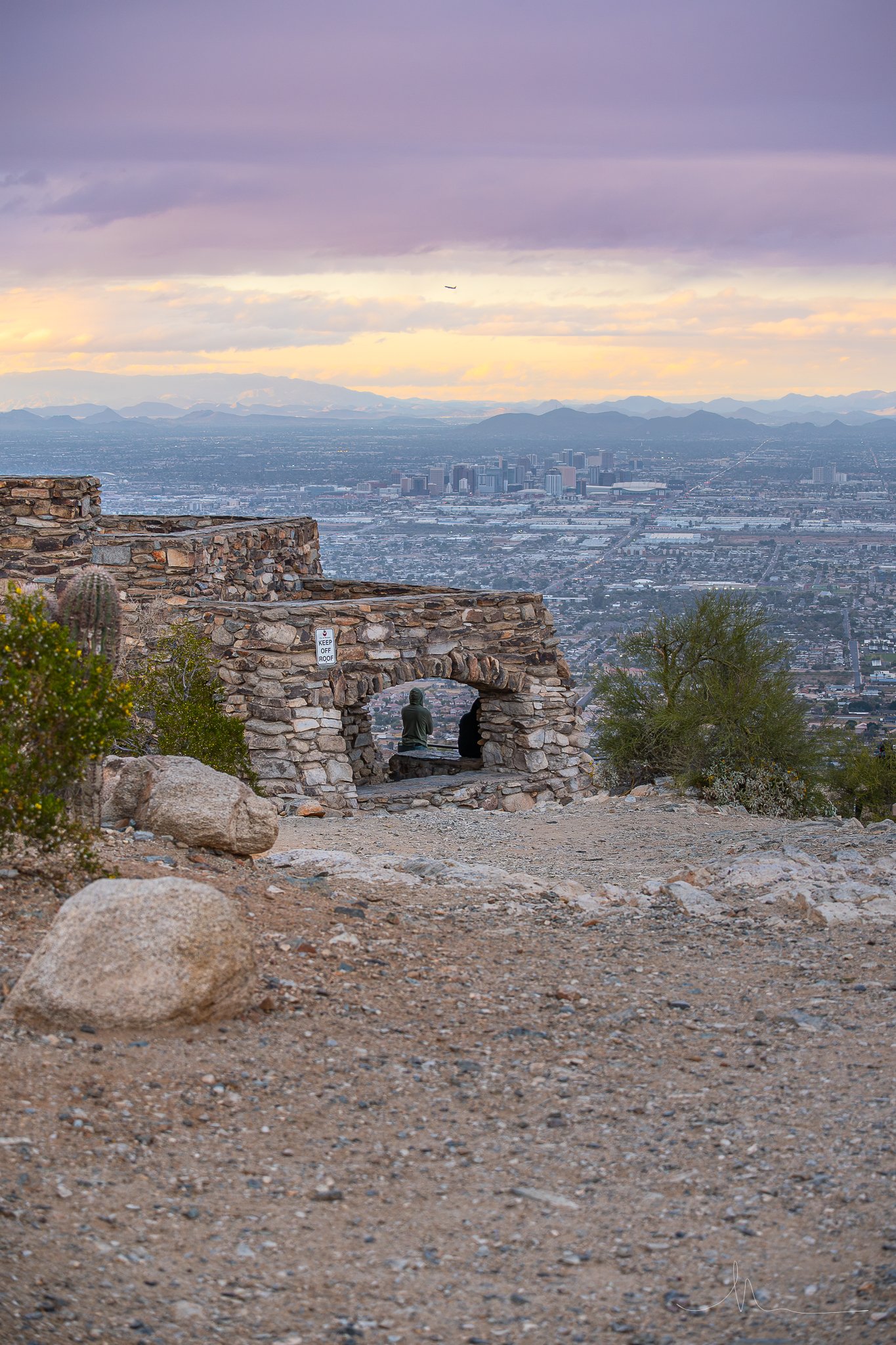 View of a cityscape from a rocky mountain lookout with stone arch structure and two statues sitting under it in the foreground, and mountains in the background at sunset.