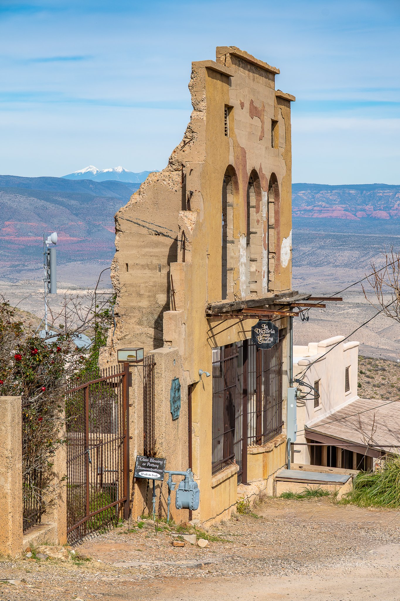 An old, partially destroyed yellow building with arched windows and a sign that reads 'The Victoria Glass Blowing Studio.' The building is on a dirt path with a fence and some machinery nearby, with mountains and snow-capped peaks in the background.