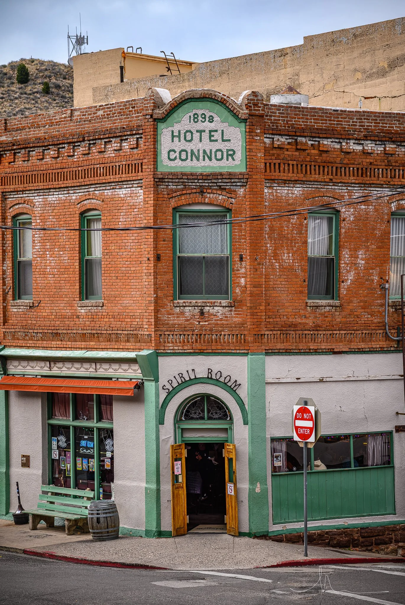 A two-story brick building with a green and white sign reading 'Hotel Connor 1898' on the top front. Below, a storefront named 'Spirit Room' with an arched entrance, green trim, and open wooden doors. A green bench, a barrel, and a 'Do Not Enter' sig