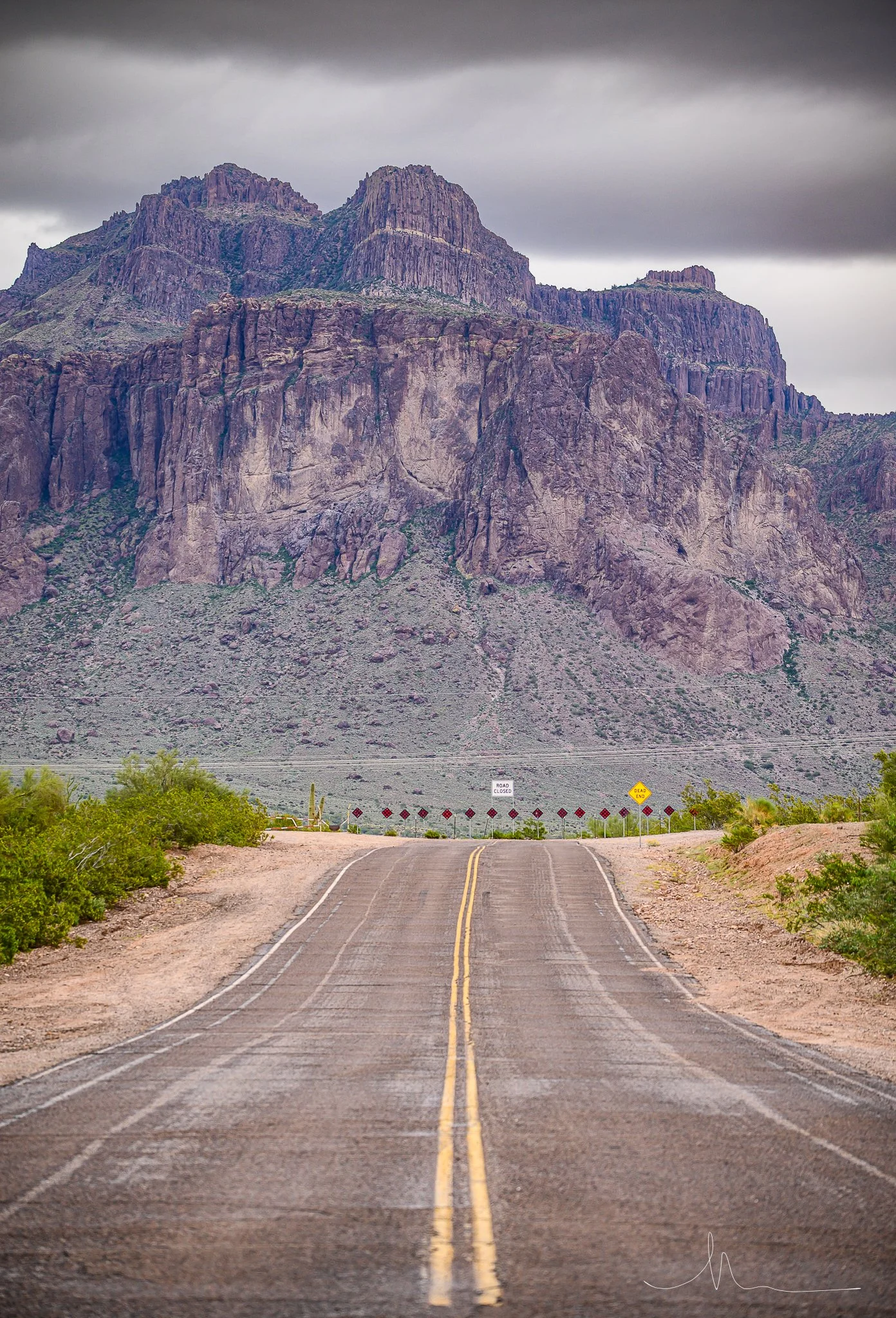 A deserted desert road with a double yellow line running through it, leading towards a large rugged mountain range under a cloudy sky.