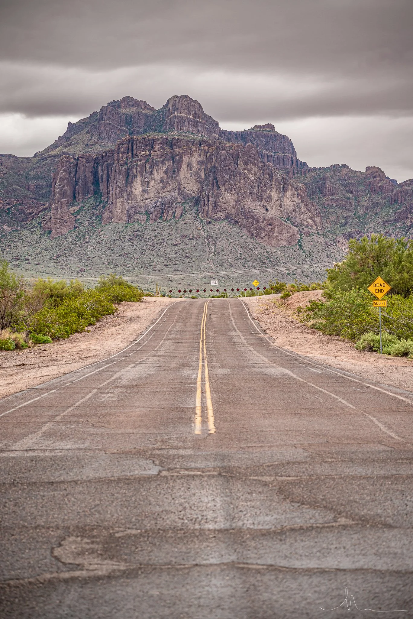 Empty desert road with yellow dividing lines, surrounded by green shrubs, leading towards large rocky mountains under cloudy sky. Sign on right reads "Dead End 500 FT".