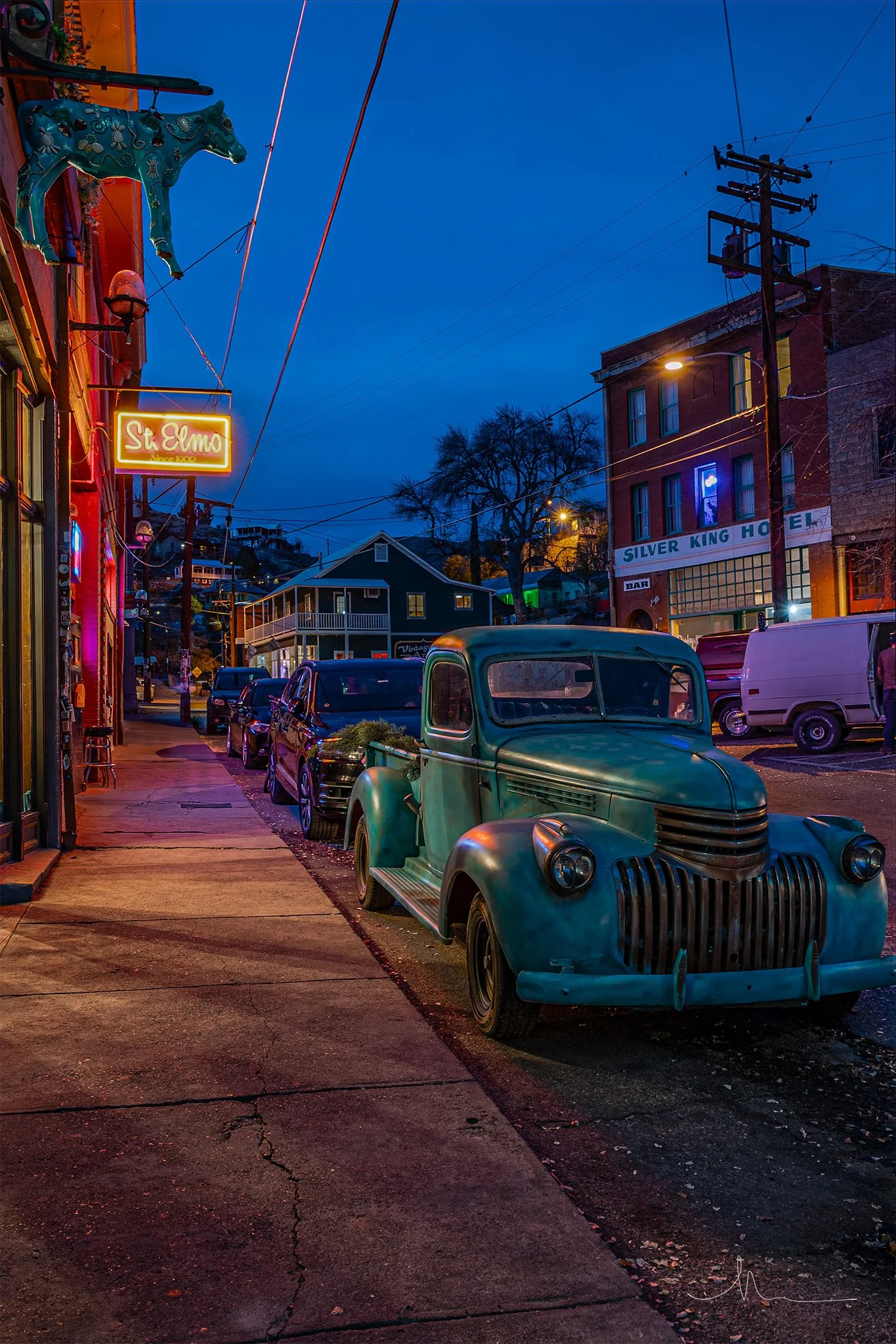 A vintage green truck parked along a city street at night, with neon signage reading 'St Elmo' and a building sign for 'Silver King Hotel.' Neon lights and streetlights illuminate the scene, and there are other parked cars along the sidewalk.