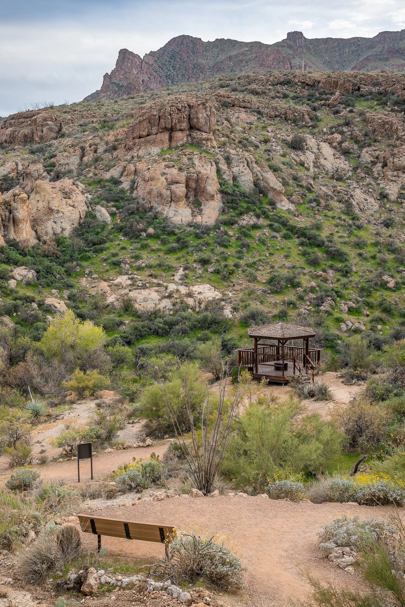 A desert landscape with rugged mountains, green shrubs, and a wooden gazebo on a dirt path.