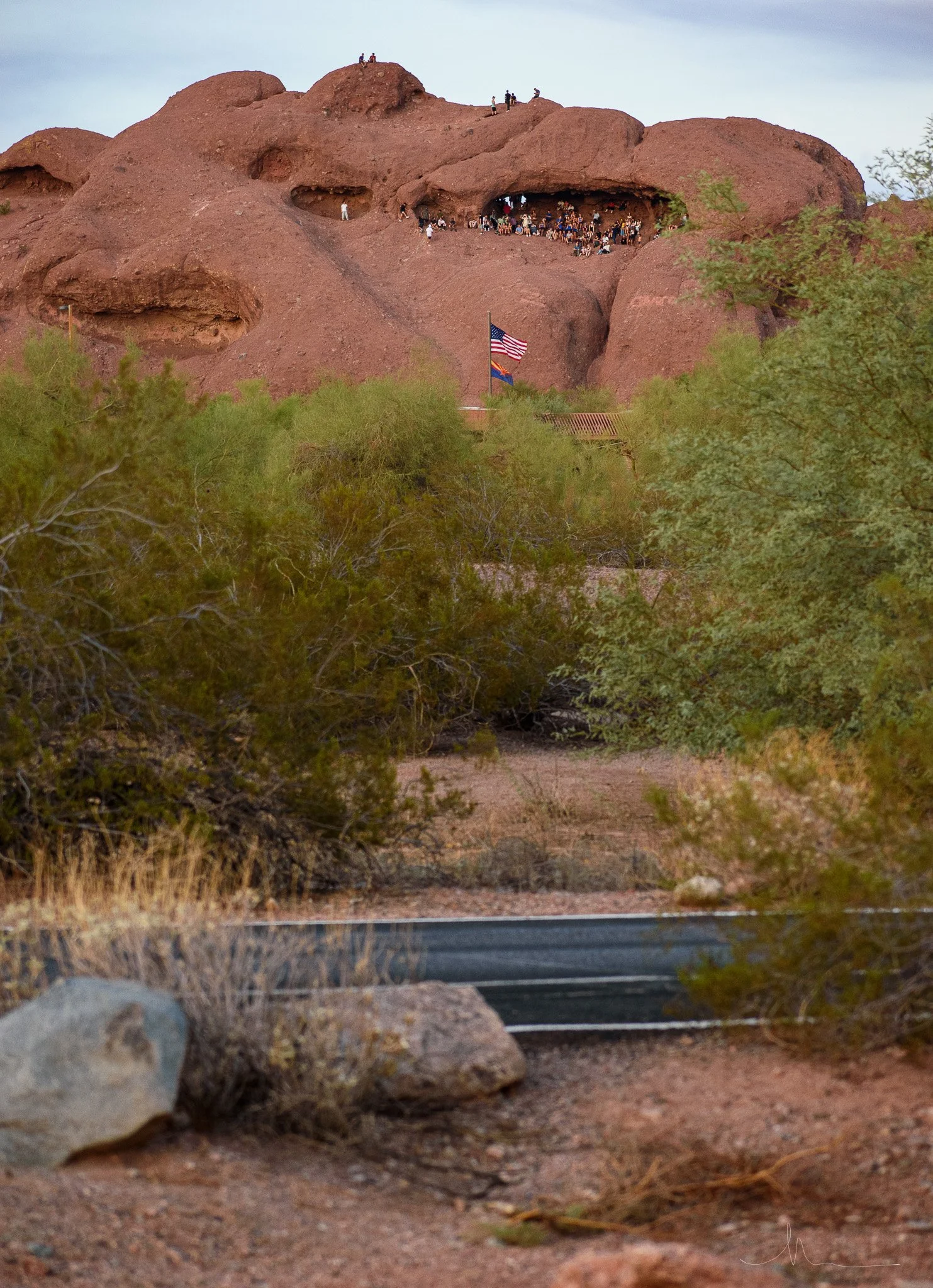 People gathered at the entrance of a large, reddish-brown rock formation with caves and ledges, with an American flag flying in the foreground. The area is desert-like with sparse greenery and bushes.