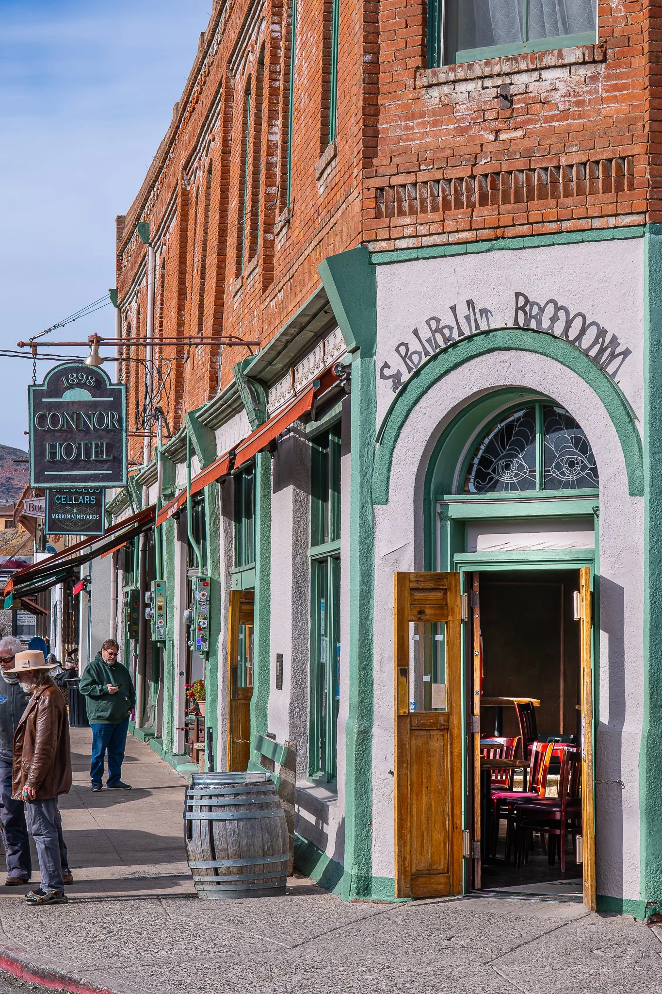 Street view showing the Connor Hotel with a hanging sign, and a storefront with a green and white exterior and open wooden door leading into a room with chairs inside. Several people are on the sidewalk.