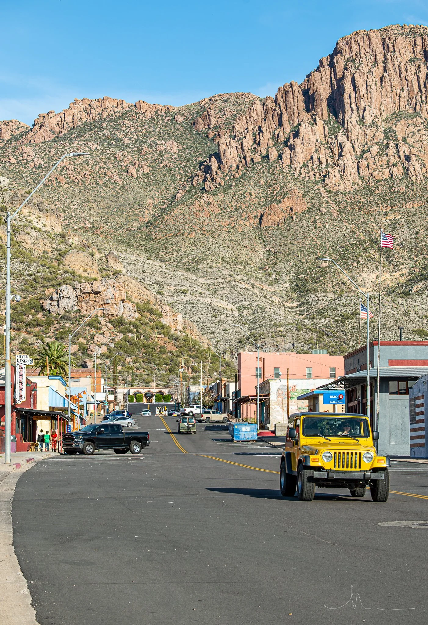 Street view of a small town at the base of rocky, desert mountains with blue sky, American flags, a yellow Jeep, cars parked along the street, and buildings with various signs.
