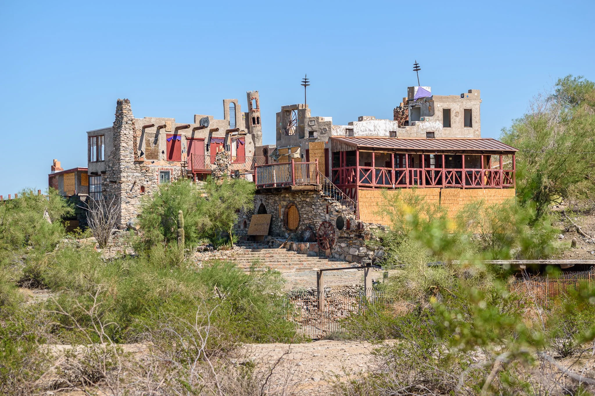 An abandoned, desert-style house with a rustic, weathered appearance, surrounded by sparse desert vegetation under a clear blue sky.
