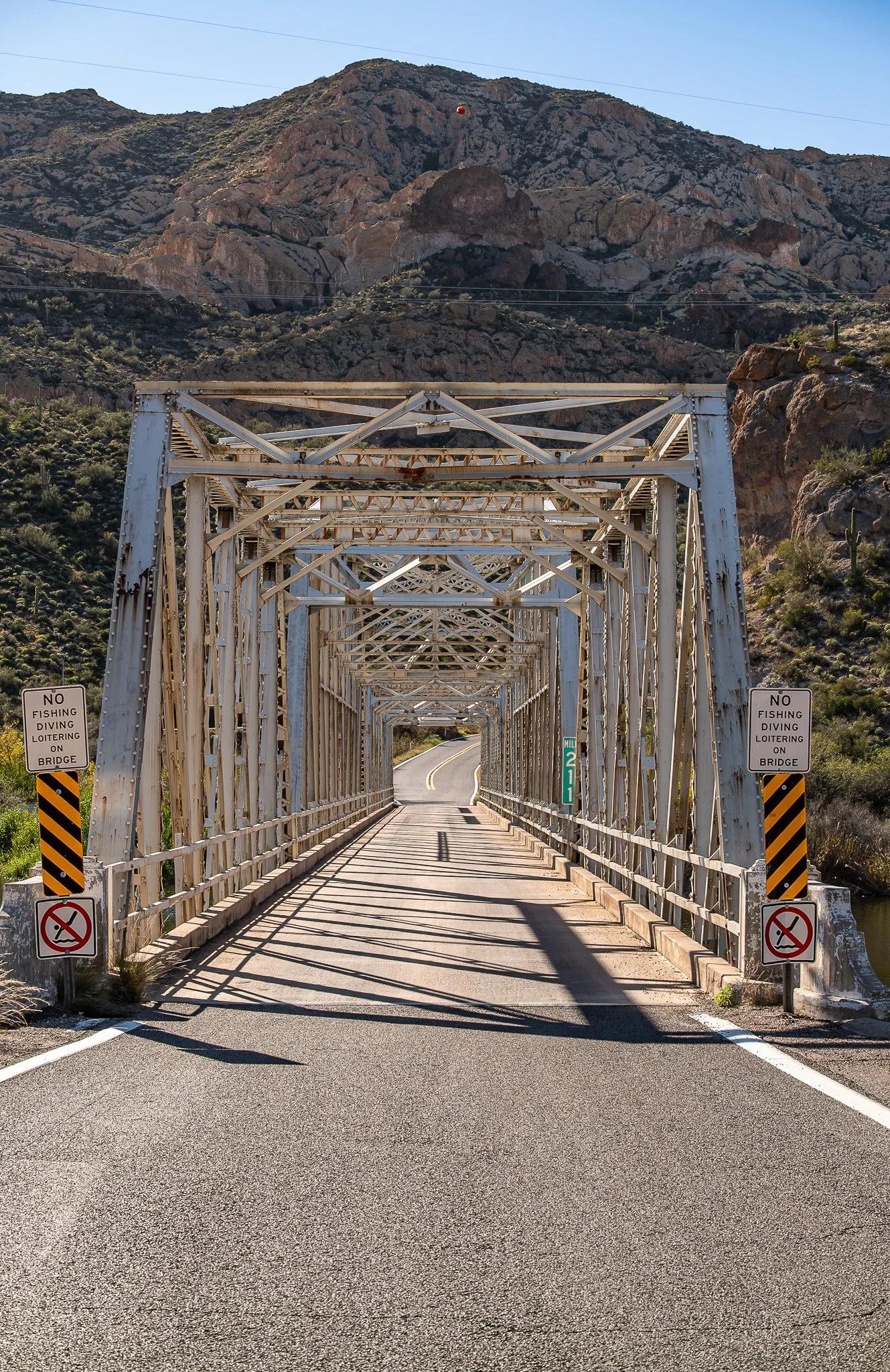 A small metal bridge over a waterway in a mountainous area, with signs indicating no fishing, diving, or loitering on the bridge.