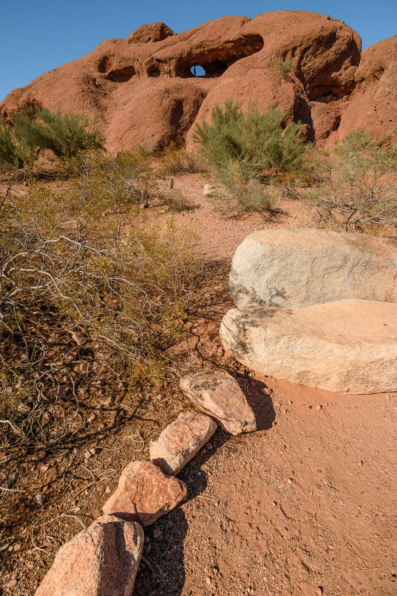 Desert landscape with reddish rocks, sparse desert bushes, and a natural rock formation with holes in it, under a clear blue sky.