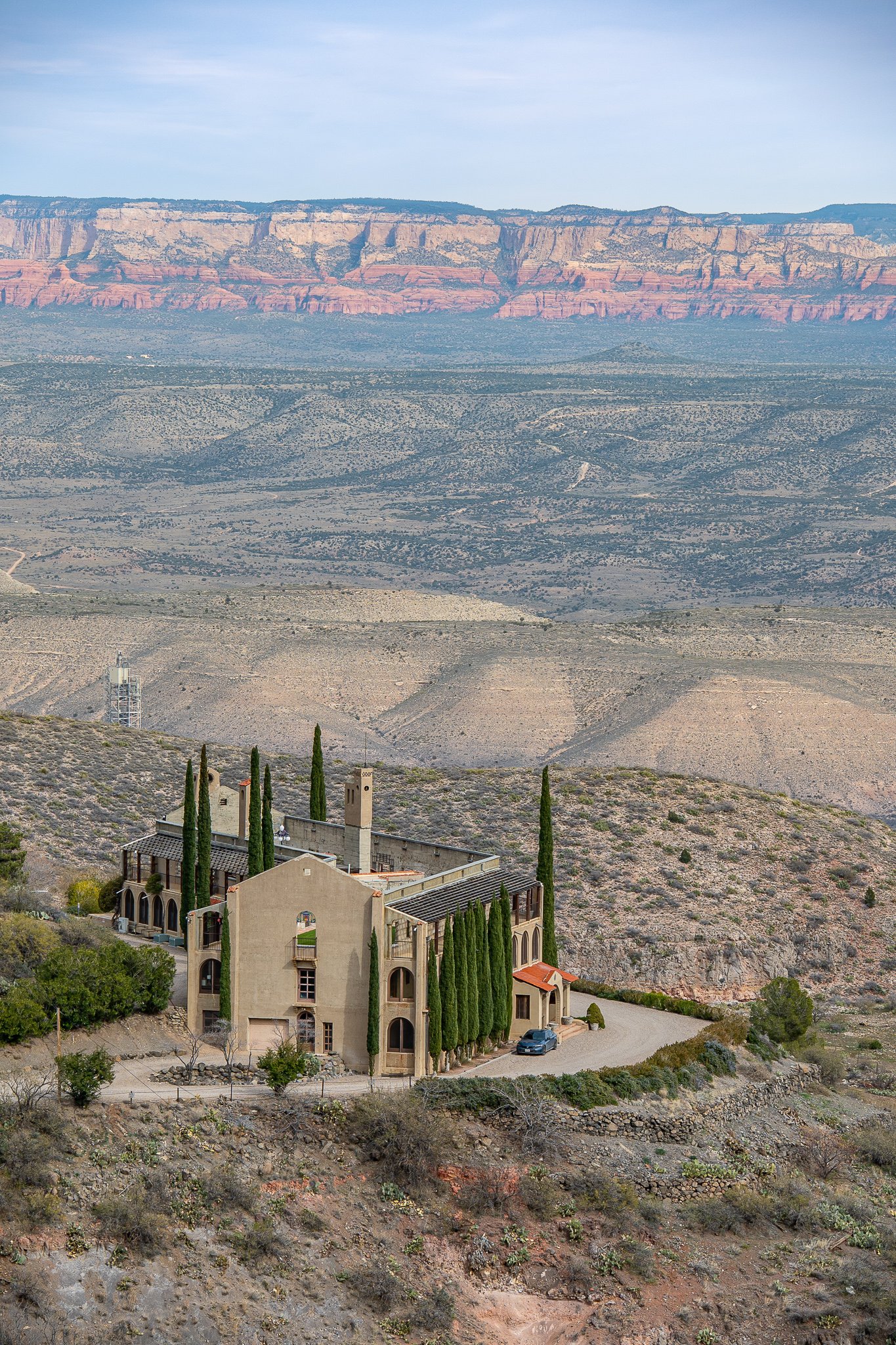 A house with tall cypress trees on a hillside in a desert landscape with layered cliffs in the background.