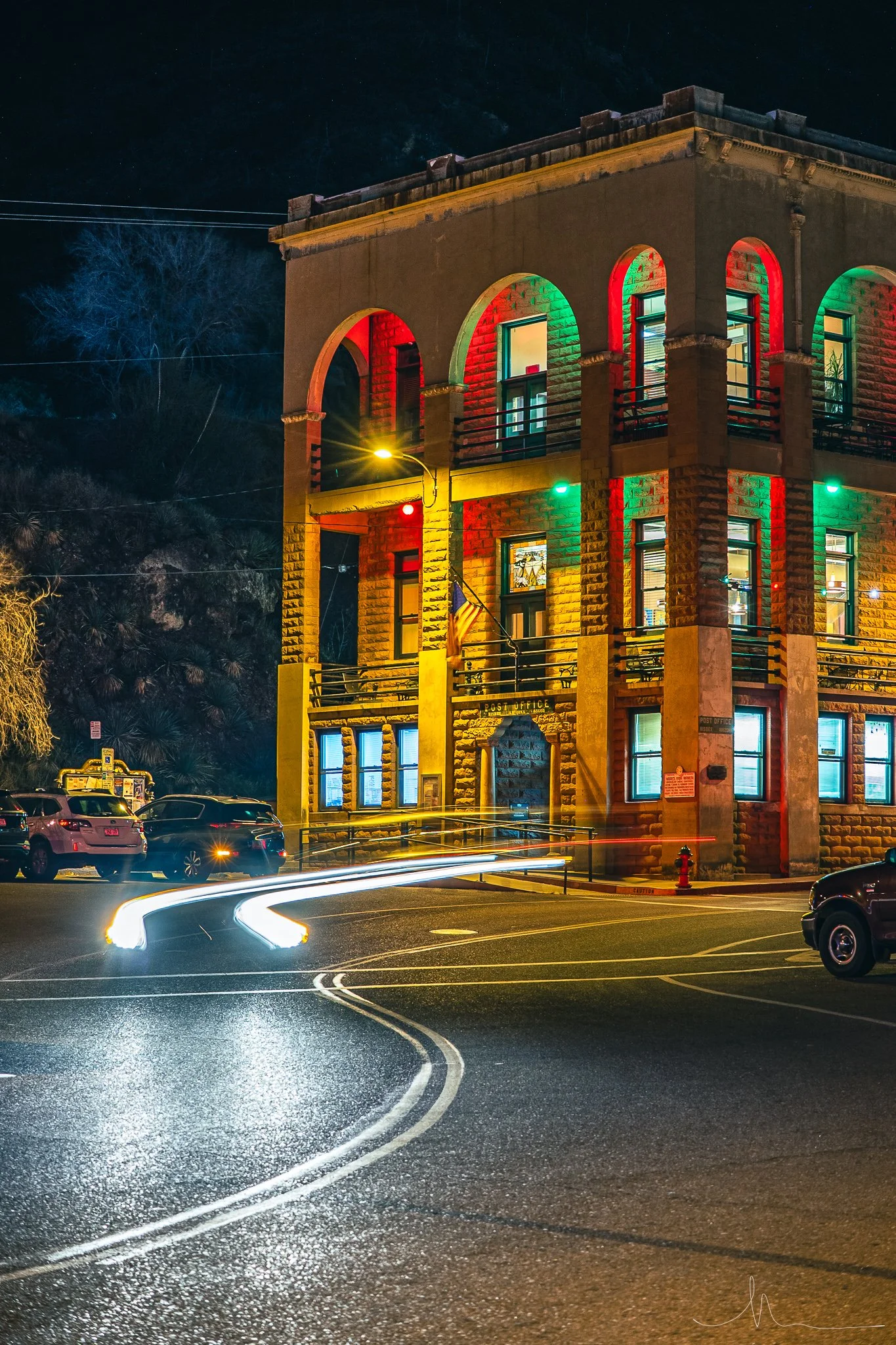 Night view of a historic stone building decorated with red, green, and blue Christmas lights. The building has arched windows and a plaque that reads 'Post Office.' Light trails from passing cars are visible on the street in front.