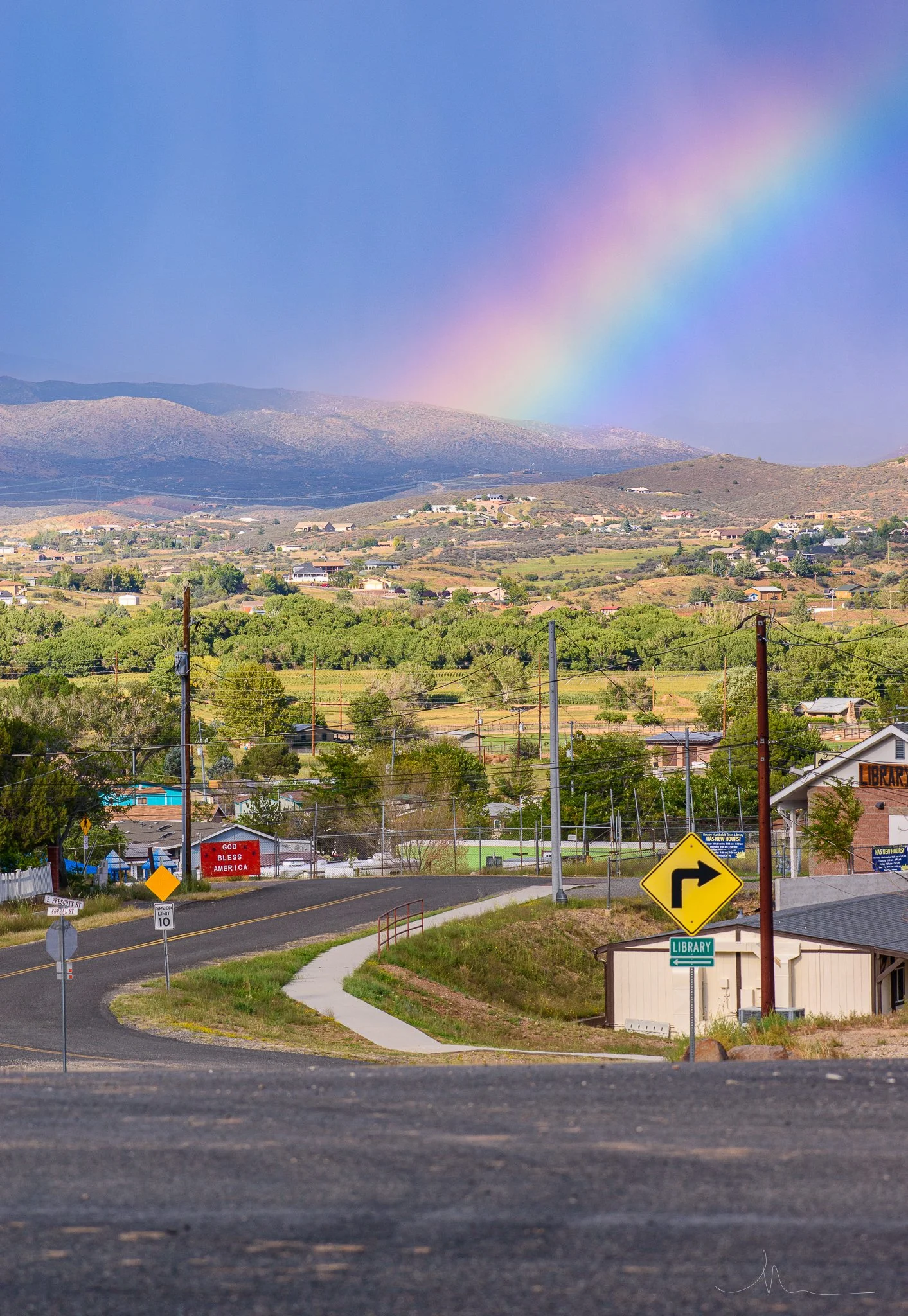 A small town near a rainbow with a blue sky and green hills in the background. There are traffic signs, a sidewalk, and a building with a sign that says 'Library' in the foreground.