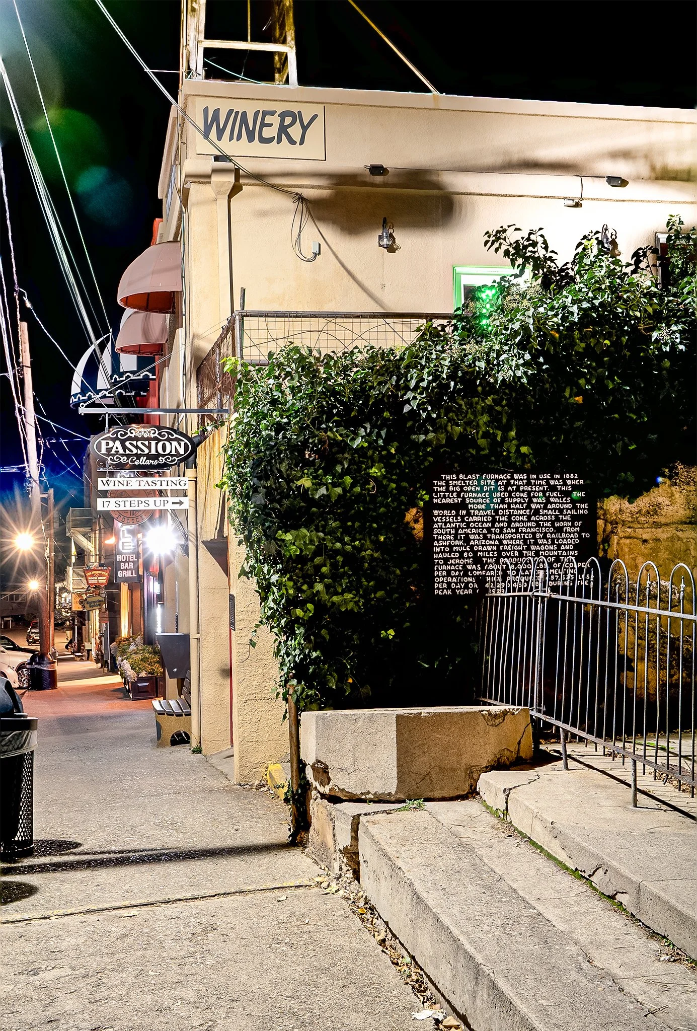 A nighttime street scene with a sign that reads 'WINERY' on a beige building. There are other signs, including one for 'PASSION Cellars' and a hotel bar grill. The street is lit by streetlights and storefront lighting, with parked cars visible on the