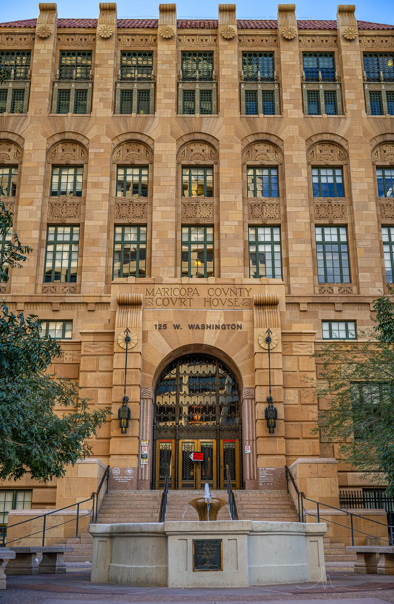 Facade of the Maricopa County Courthouse, a tall historic building with stone architecture, multiple windows, and an arched entrance at 125 W. Washington, with a small fountain in front.