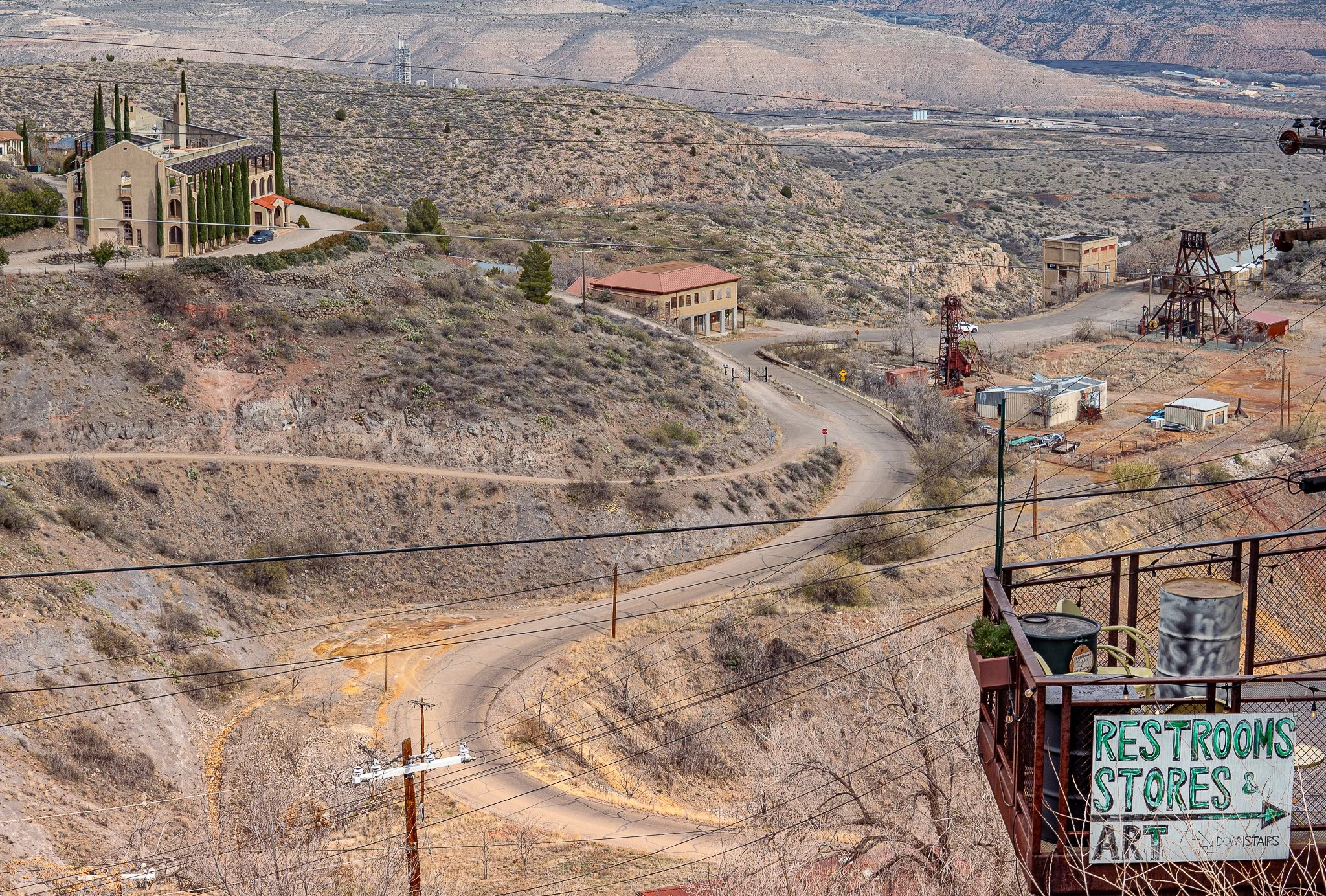 Scenic view of a hilly, semi-arid landscape with dirt roads, sparse vegetation, and buildings including a church, a small store, and some industrial structures, with power lines and a sign pointing to restrooms, stores, and art.