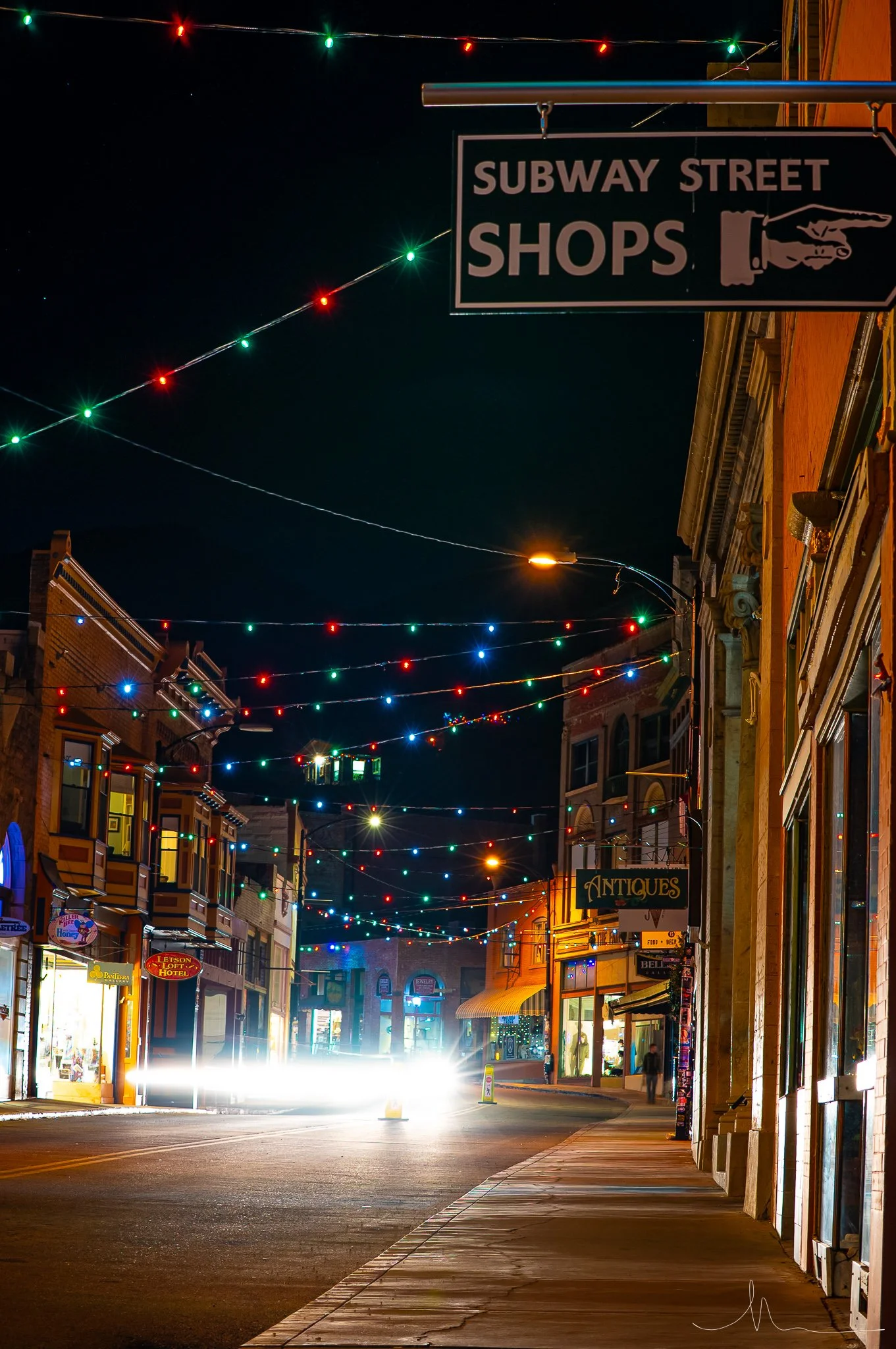 Nighttime street scene with colorful string lights, storefronts including an antiques shop, and bright headlights of an approaching vehicle.