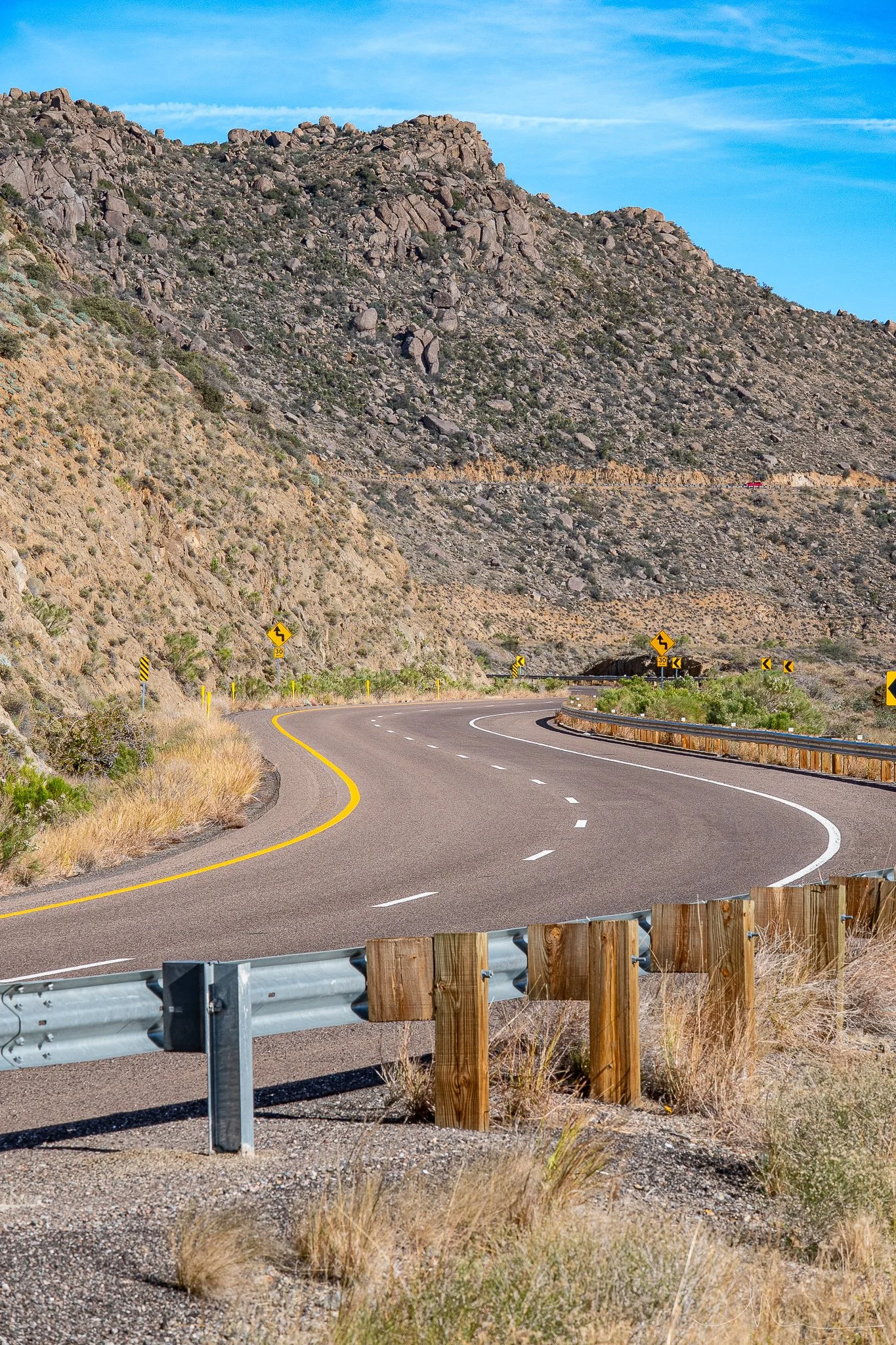 Winding mountain road with guardrails, yellow and black road signs, rocky hills, and clear blue sky.