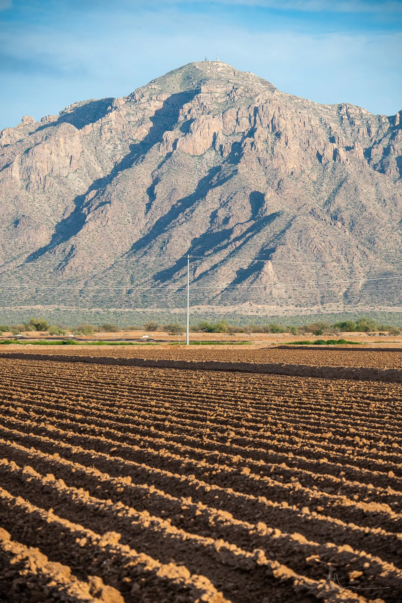 A mountain in the background with a shadow cast across its face, a utility pole in the middle ground, and freshly plowed farmland in the foreground.