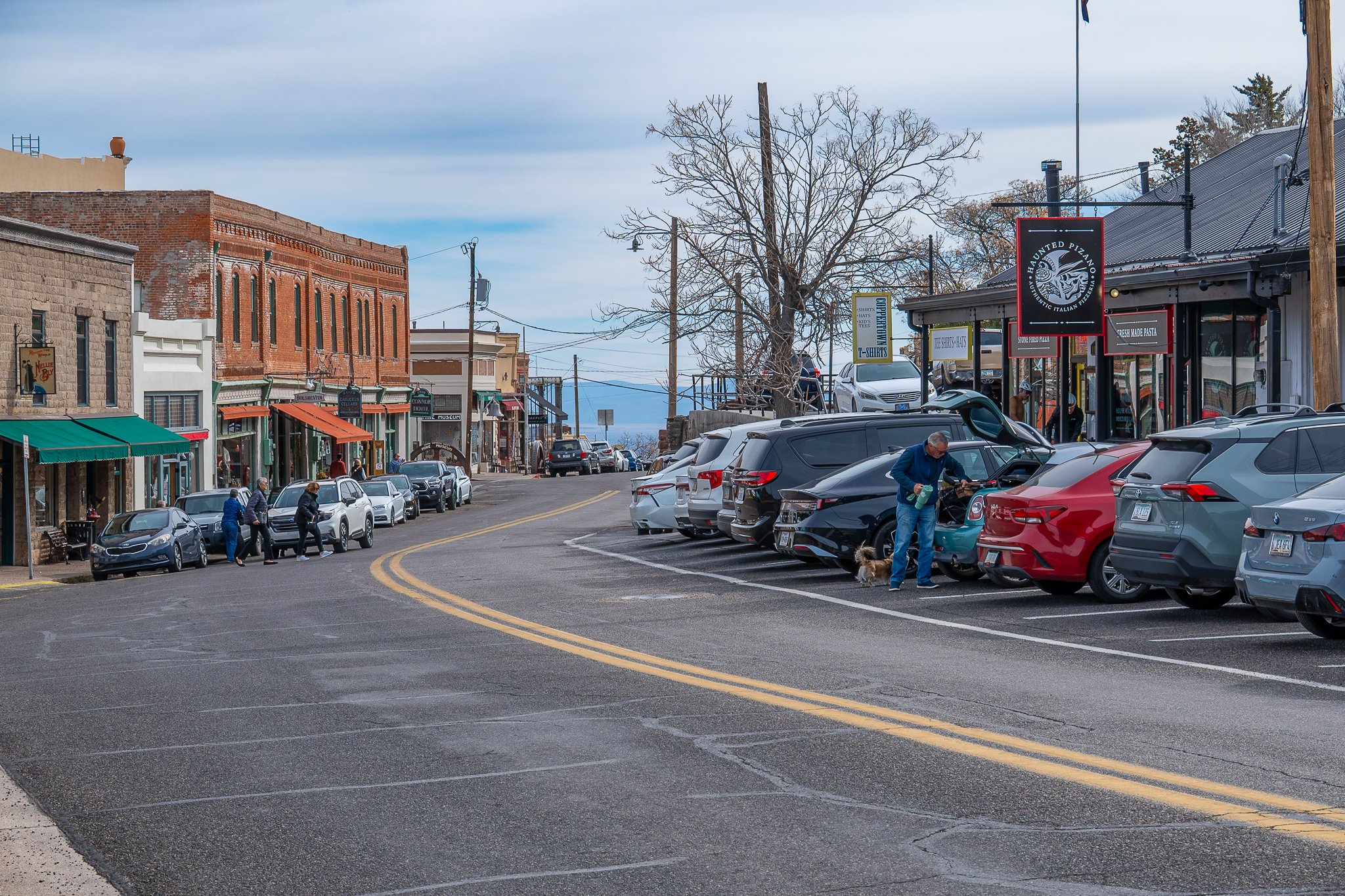 A small town street with parked cars on both sides, storefronts with awnings, pedestrians walking, and a person with a dog near a turquoise car.