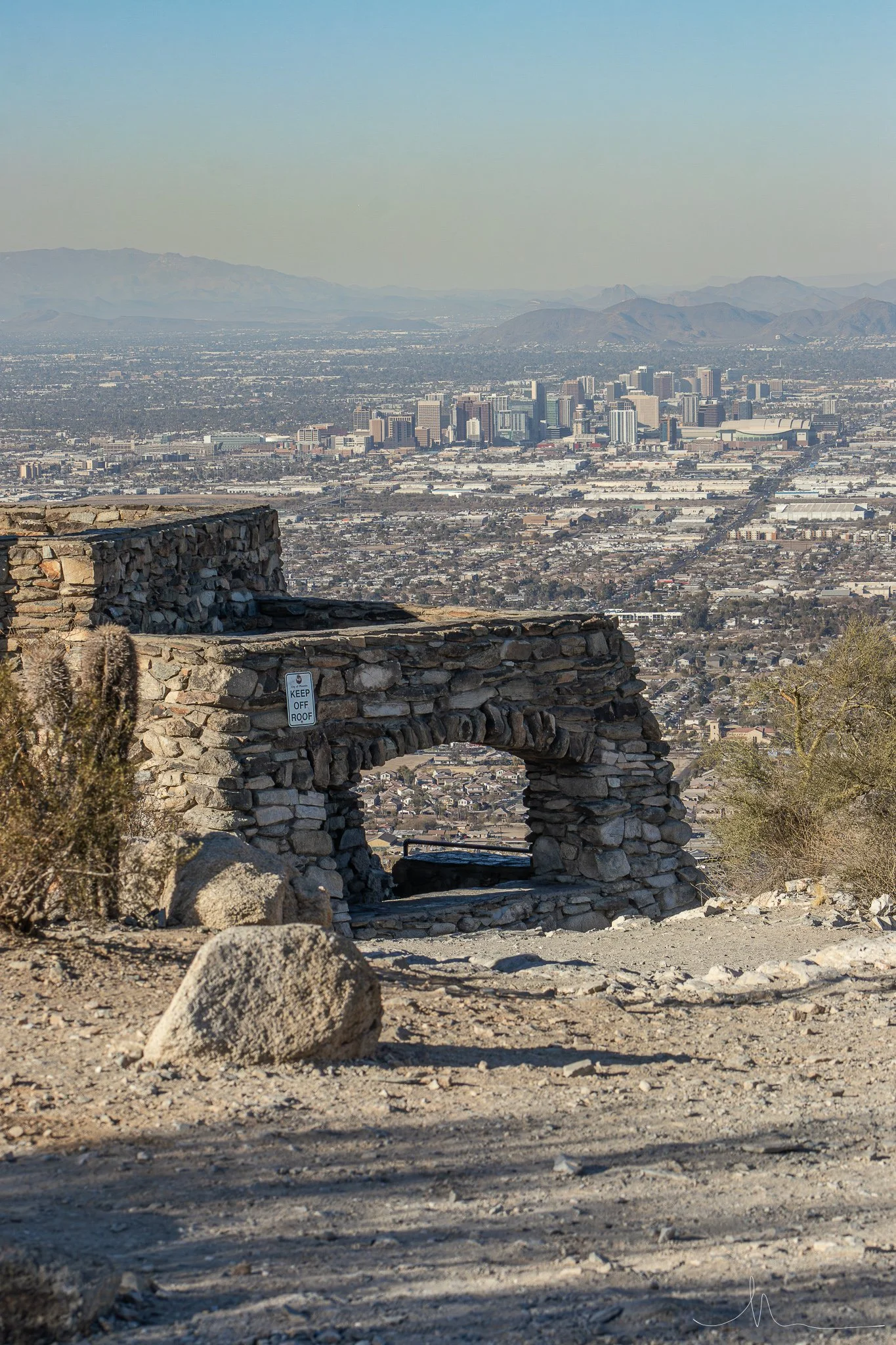 A stone arch structure on a hill with a cityscape in the background, under a clear blue sky.