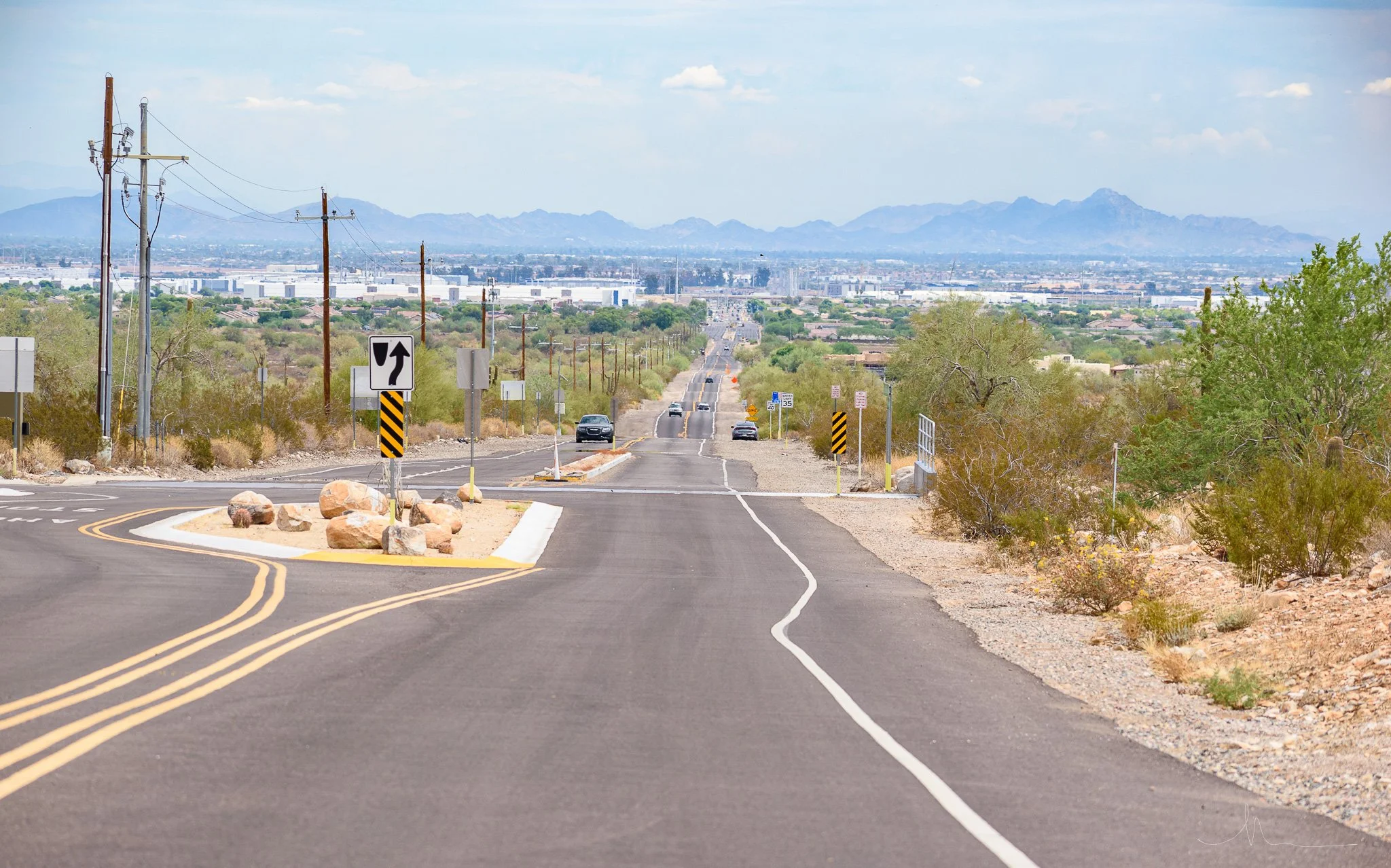 A desert road with a traffic island in the middle, chevron signs, utility poles, and mountains in the distance under a partly cloudy sky.