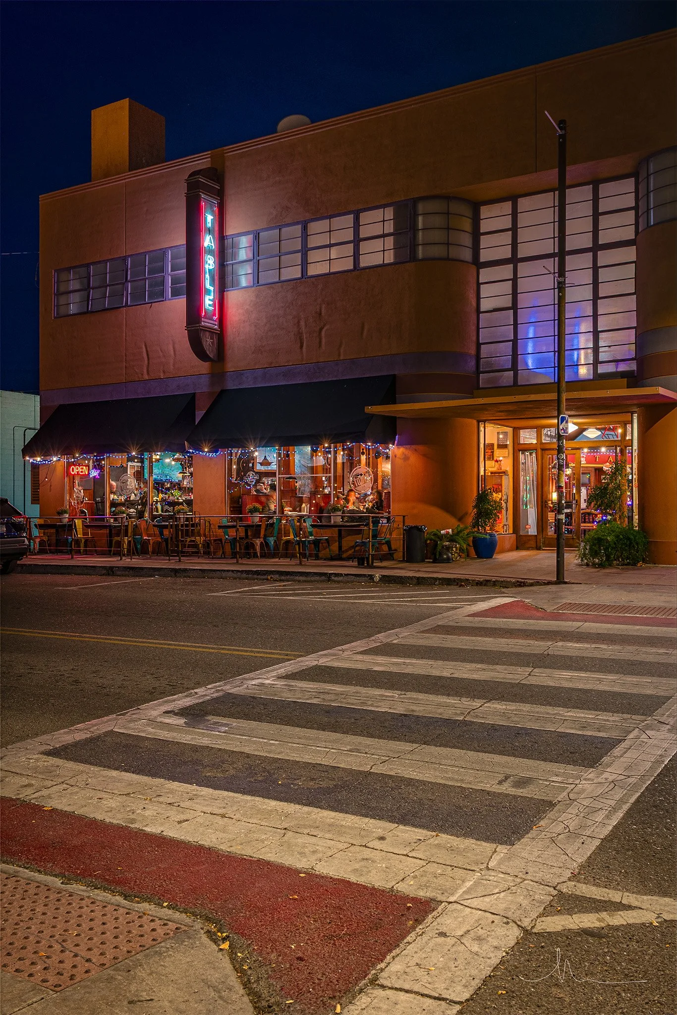 Nighttime view of a corner building with a neon sign that reads "TAPS". The building has large windows with interior lighting, outdoor seating, and a small street with a pedestrian crosswalk in the foreground.