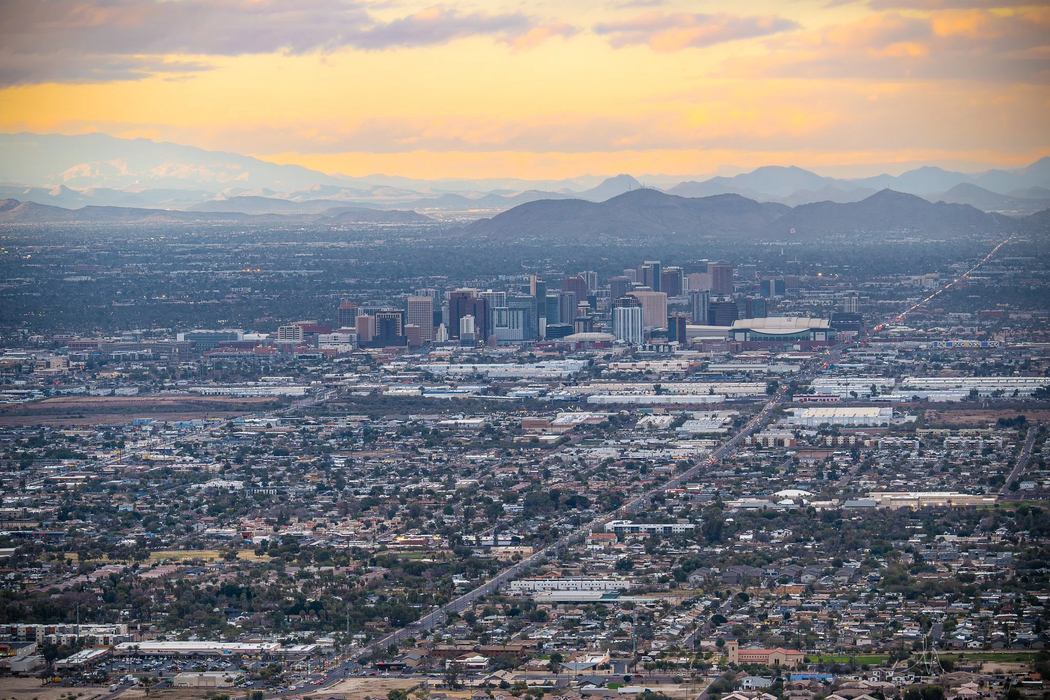 Aerial view of a large city with numerous buildings, high-rises, and a grid-like street layout, with mountains in the background and a colorful sky during sunset.
