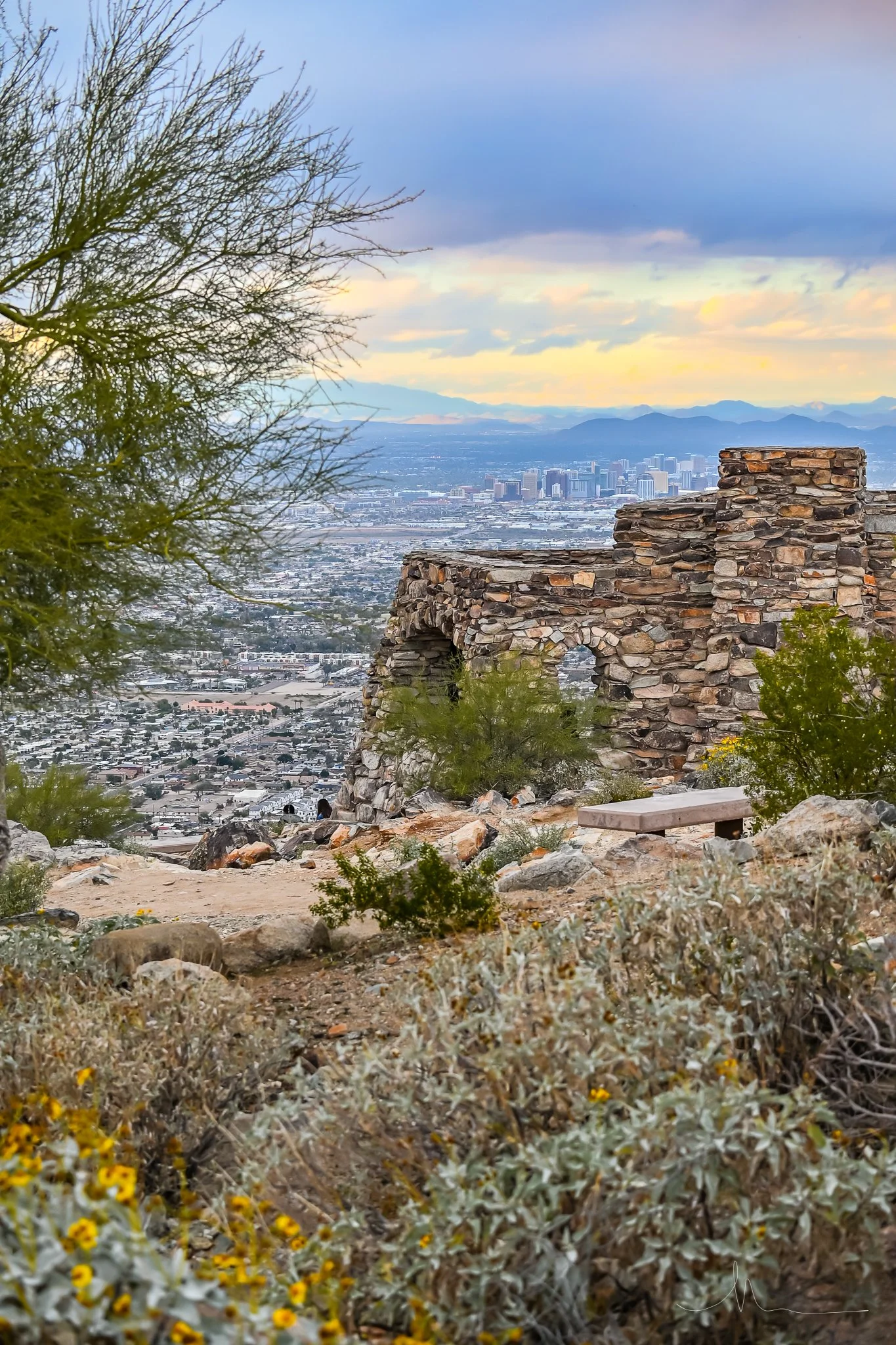 A scenic view from a mountain overlooks a city with tall buildings in the distance, featuring a stone structure with arches and desert vegetation in the foreground, with mountains and a cloudy sky in the background.