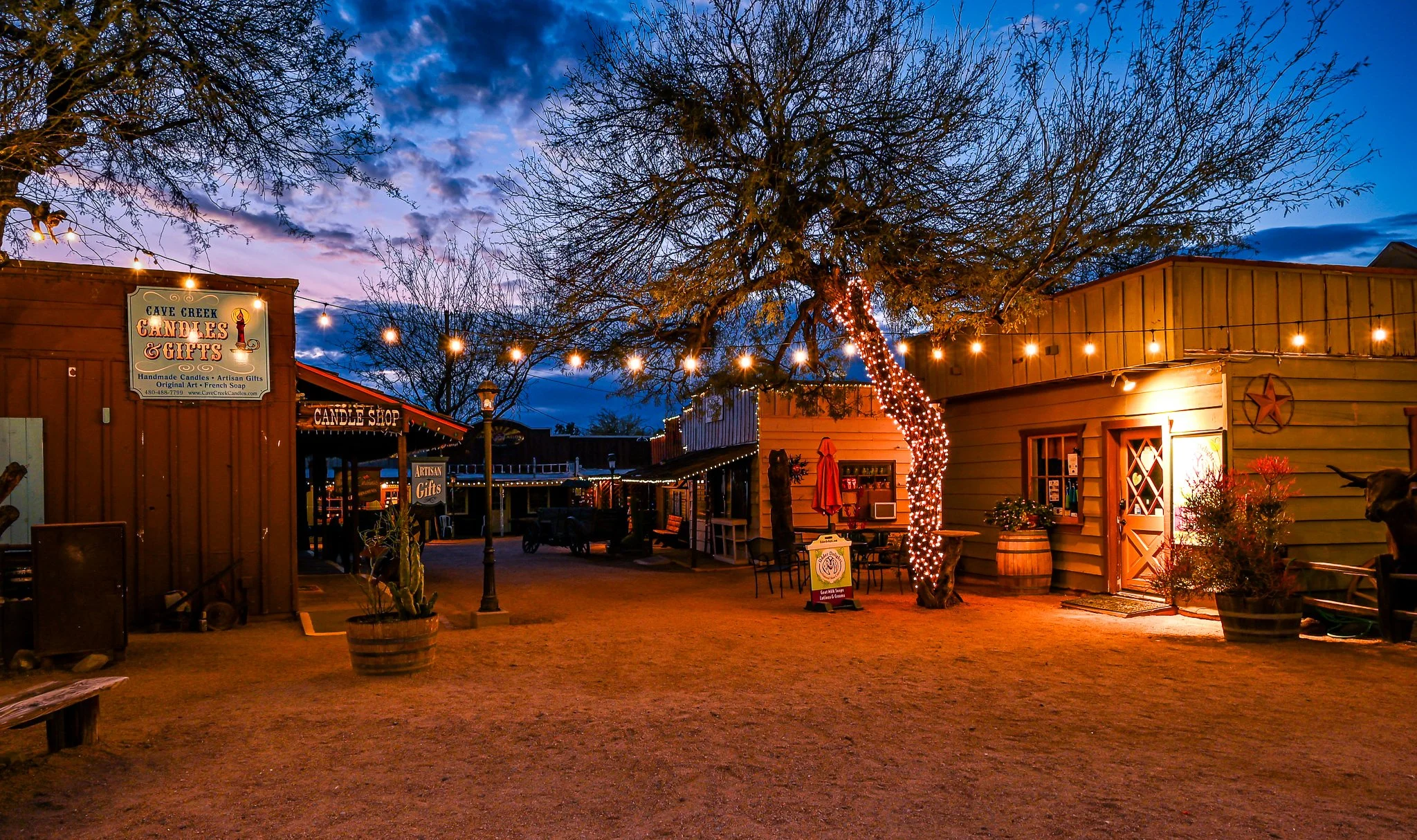 An outdoor scene at dusk with rustic buildings, string lights, a decorated tree with lights, and signs for a candle shop and artisan gifts.