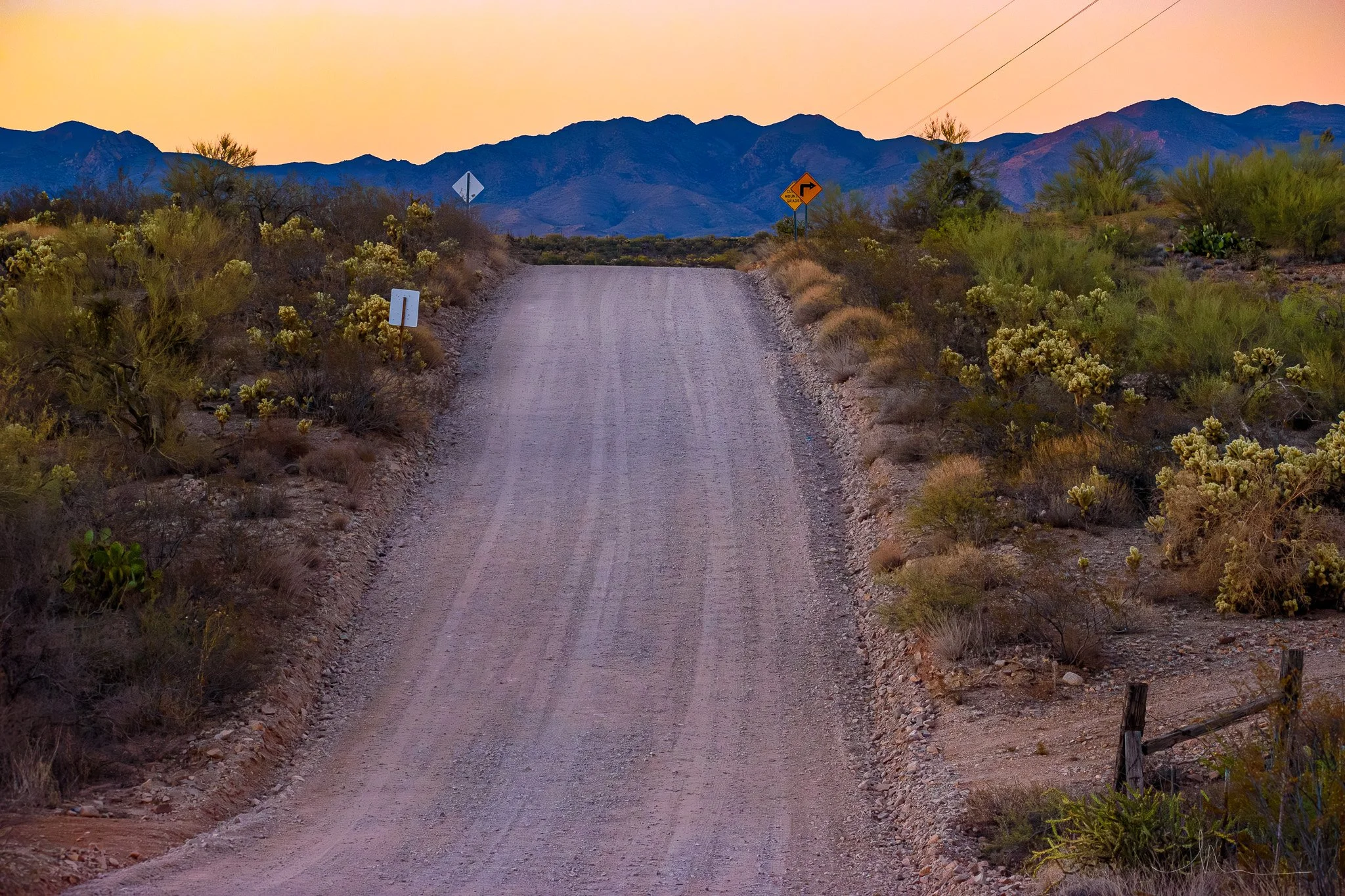 A dirt road in a desert landscape during sunset, with mountain range in the background, and several road signs along the road.