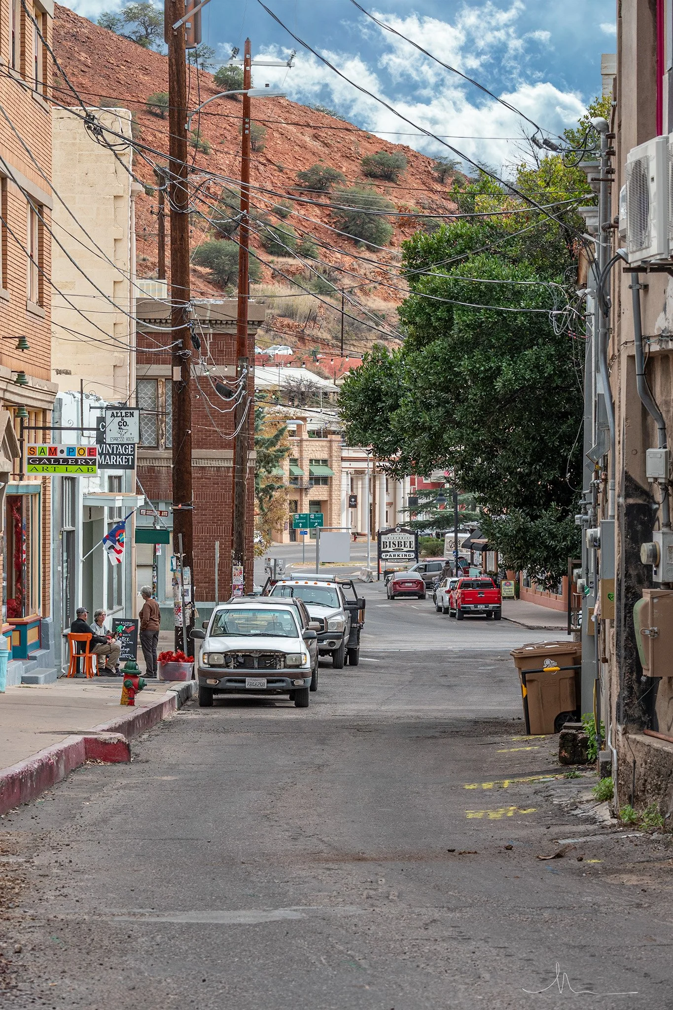 A back alley street scene in a historic small town, with parked cars, storefronts, power lines, and a hillside in the background.