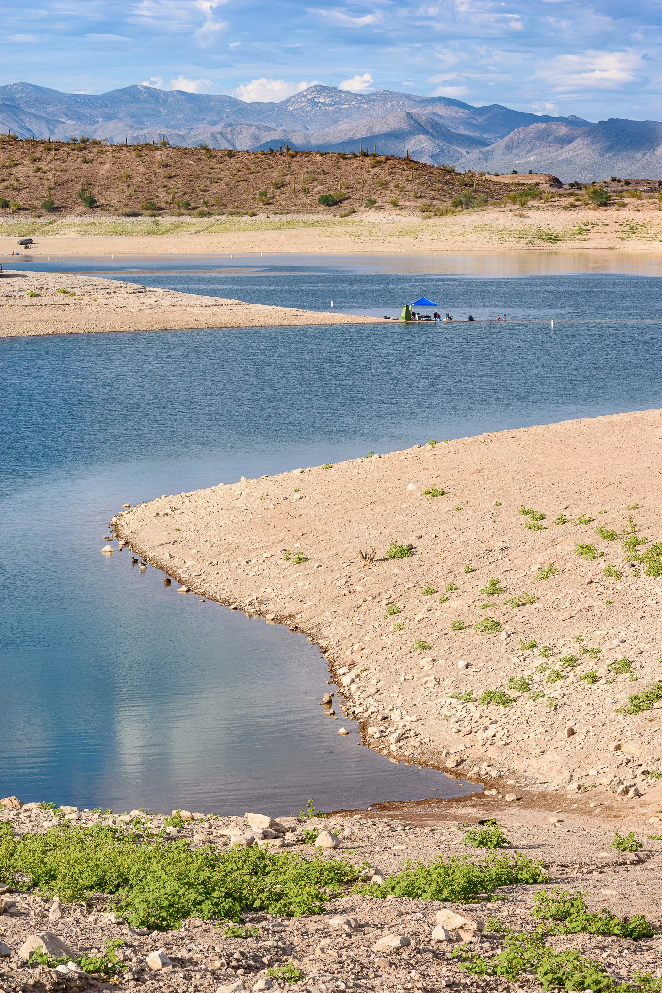 A landscape with a lake, desert terrain, mountains in the background, and a blue sky with clouds.