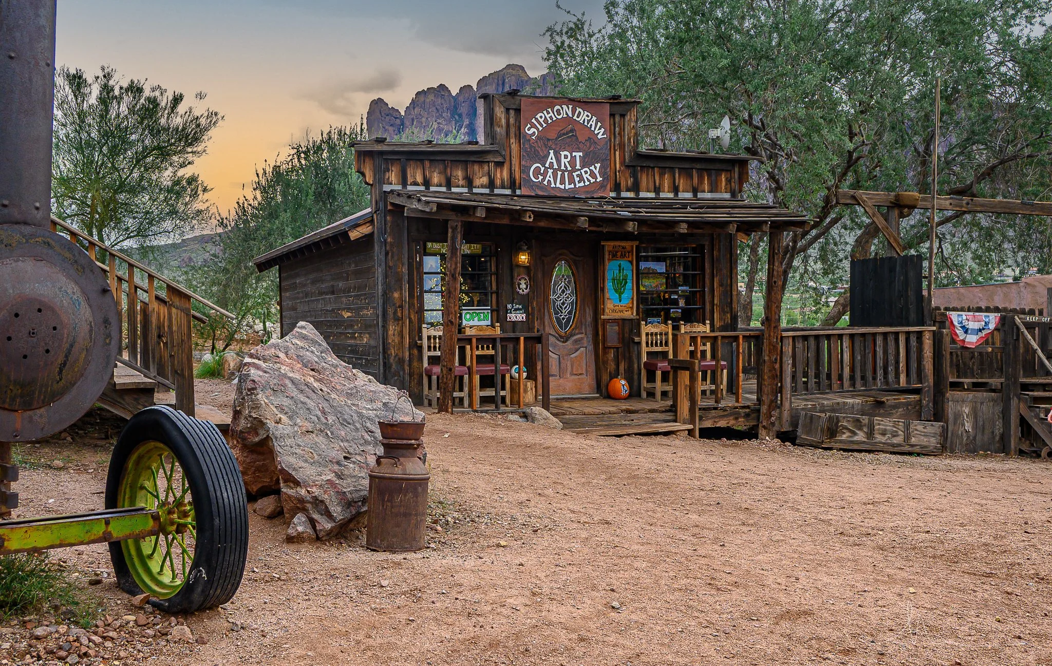 A rustic wooden art gallery building with a sign reading 'Siphon Draw Art Gallery' in a desert setting. The building is surrounded by trees, rocks, and vintage farm equipment, including a wheel and an old milk can. The sky is at sunset or sunrise, ca