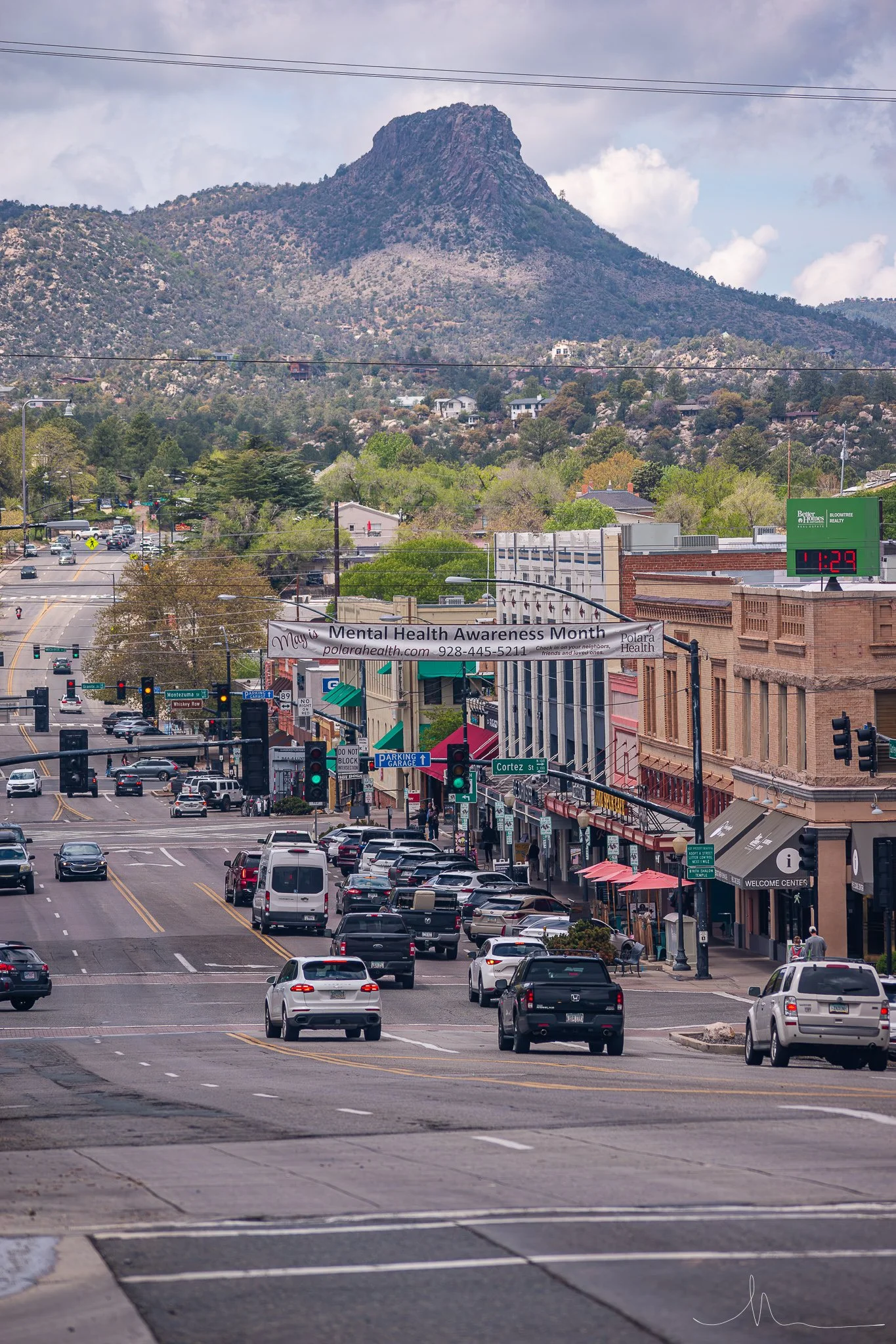 A busy downtown street with cars and pedestrians, green traffic lights, storefronts, a banner for Mental Health Awareness Month, and a scenic mountain in the background under a partly cloudy sky.