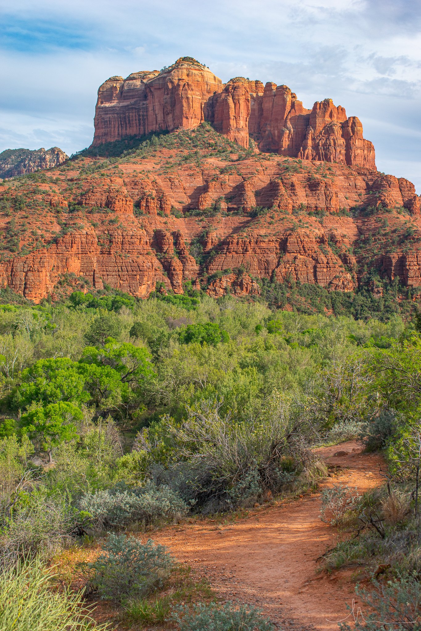 Photo of red rocky cliffs and mesas in a desert landscape with green foliage in the foreground and a partly cloudy sky.