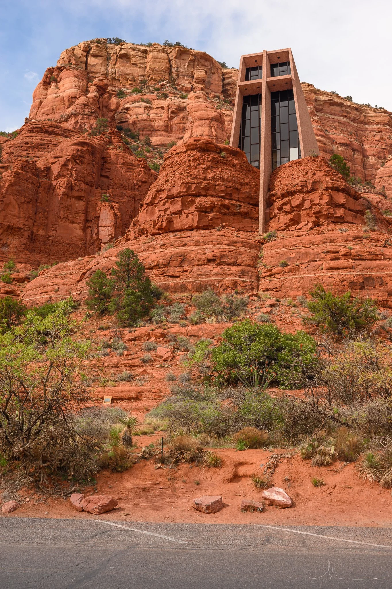 Modern building structure embedded in red rock cliffs, with sparse desert vegetation at the base and a paved road in the foreground.