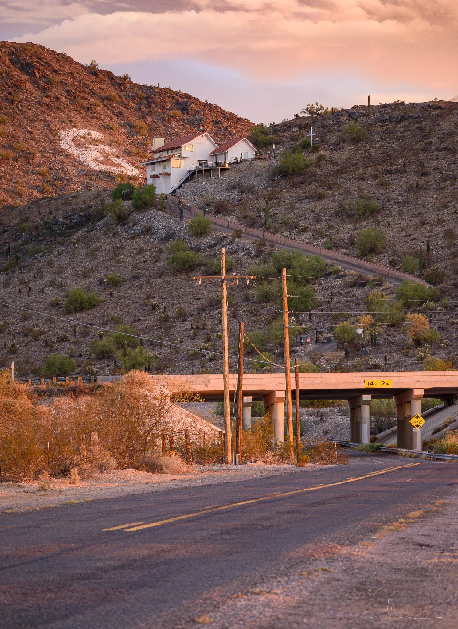 A house built on a hillside with a winding uphill driveway leading to it, a bridge with a height clearance sign, and utility poles along a road in a desert landscape during sunset.