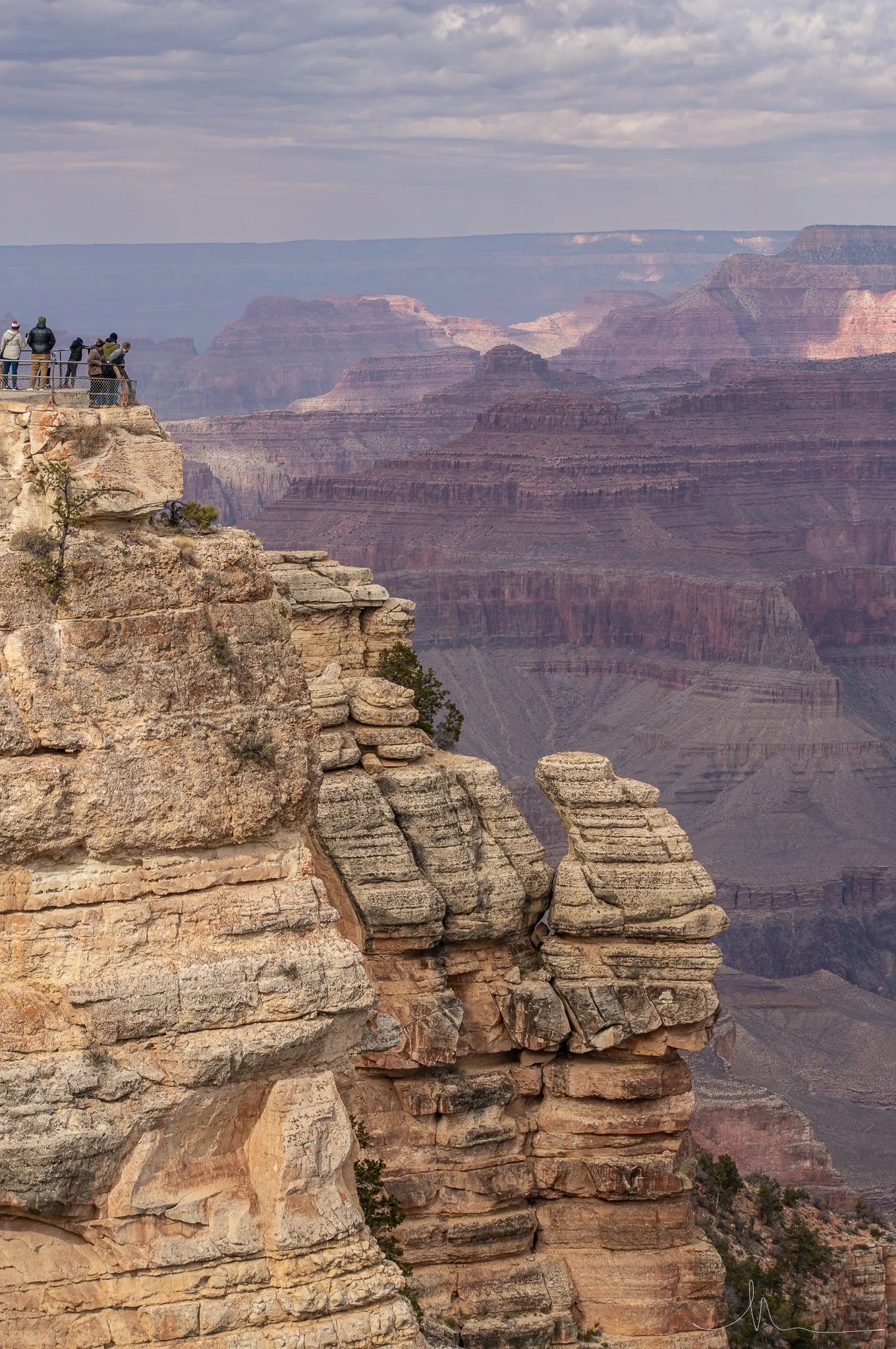 View of the Grand Canyon with a group of people on a viewing platform on the left side, overlooking layered rock formations and deep canyon valleys under a cloudy sky.