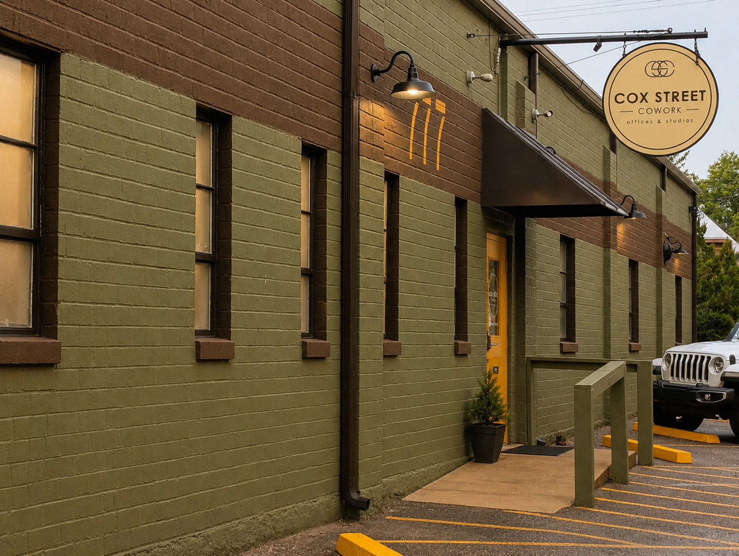 Exterior of a building with a green and brown brick wall and a sign for Cox Street Cowork. The sign is round, beige with black text. There are black outdoor lights on the wall, a neon sign of a beer mug, a yellow door, a parking area with yellow park