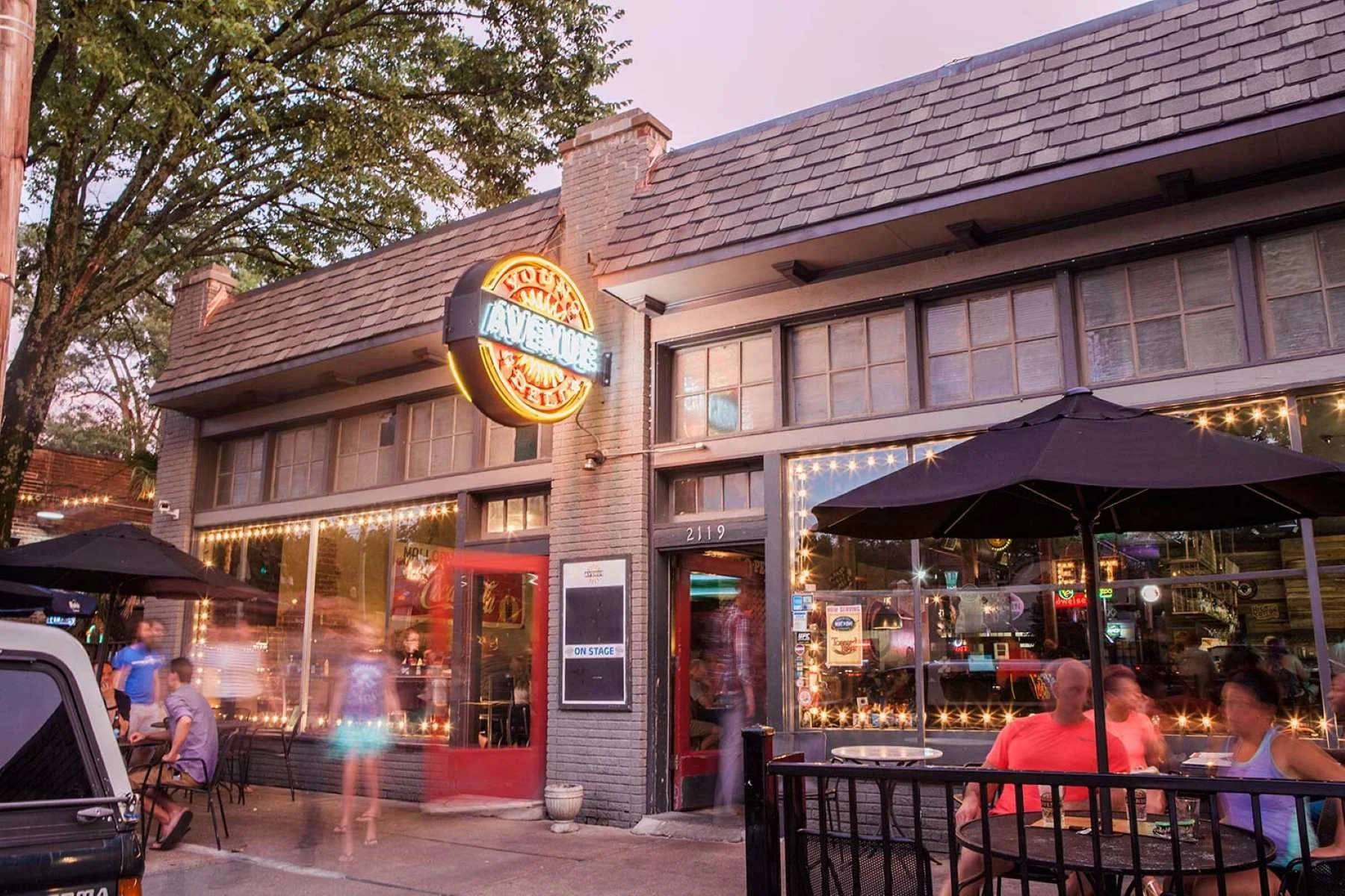 A lively restaurant with an outdoor patio at dusk. The building has large glass windows and a neon sign that reads 'You' and 'Famous'. There are string lights illuminating the patio area, with people sitting at tables under umbrellas and some walking by. The scene has a cozy, inviting atmosphere.