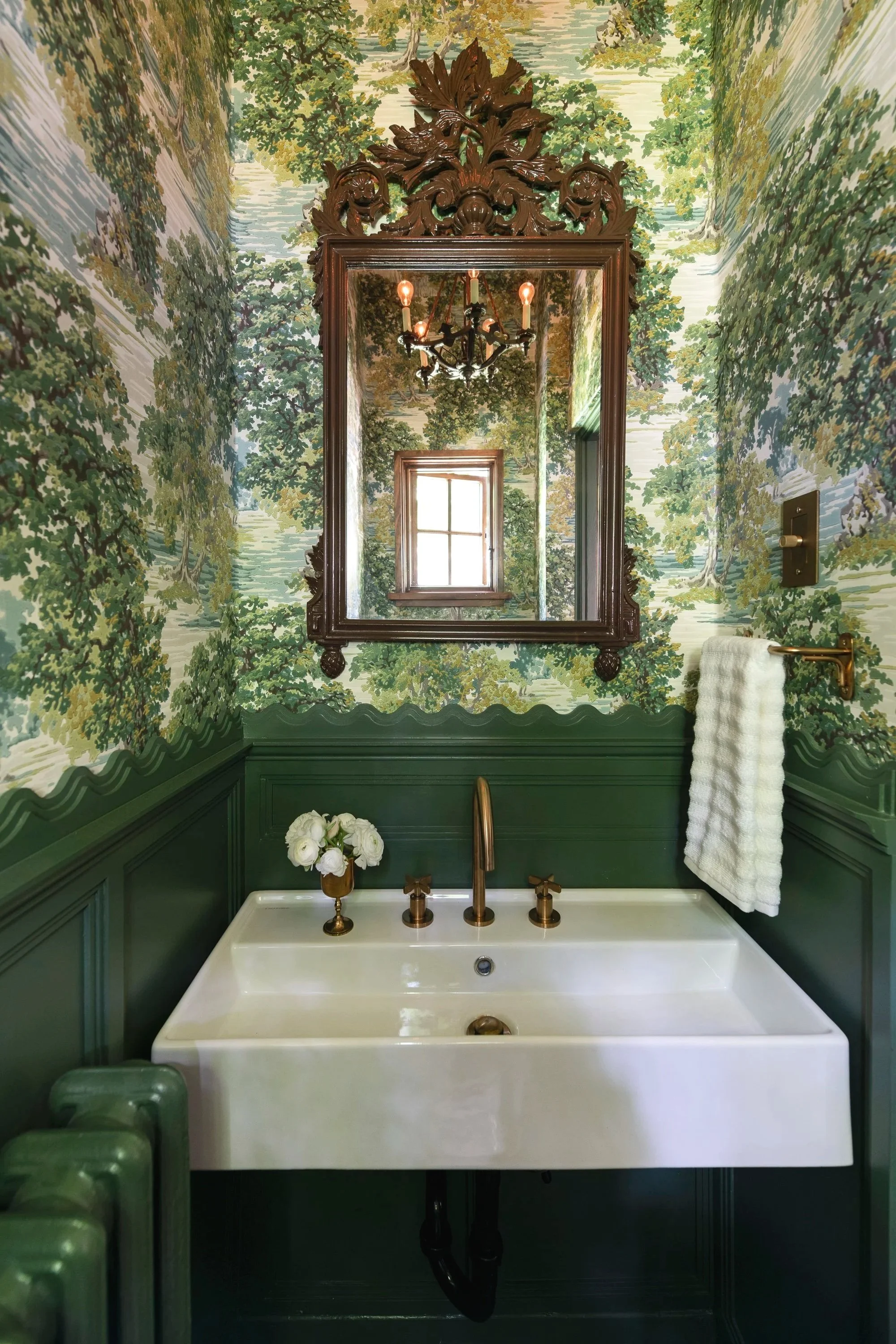 A bathroom with a large white bathtub and green paneling. The wall is decorated with wallpaper depicting a lush green forest scene. A brass towel holder with a rolled white towel is mounted on the wall. Above the tub, a large, ornate wooden-framed mi