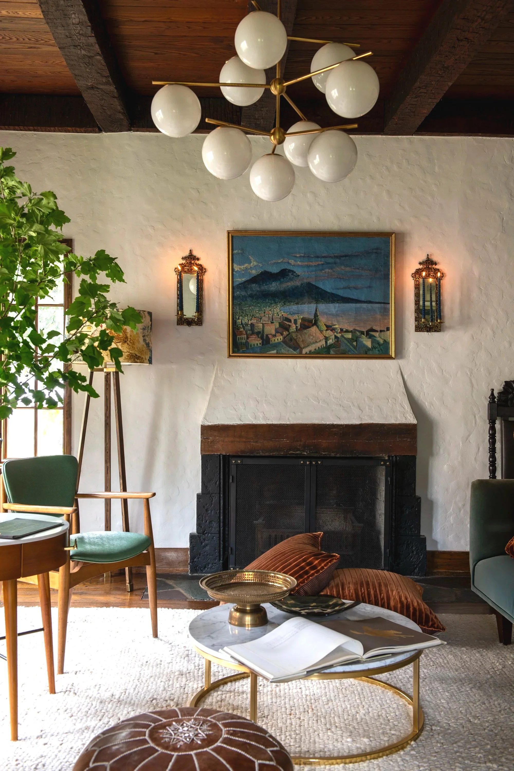 Living room with a white textured wall, a black brick fireplace, and a painting of a mountain scene above the mantel. Decor includes two ornate wall sconces with candles, a tall lamp with a floral shade, and a modern chandelier with white globes and 