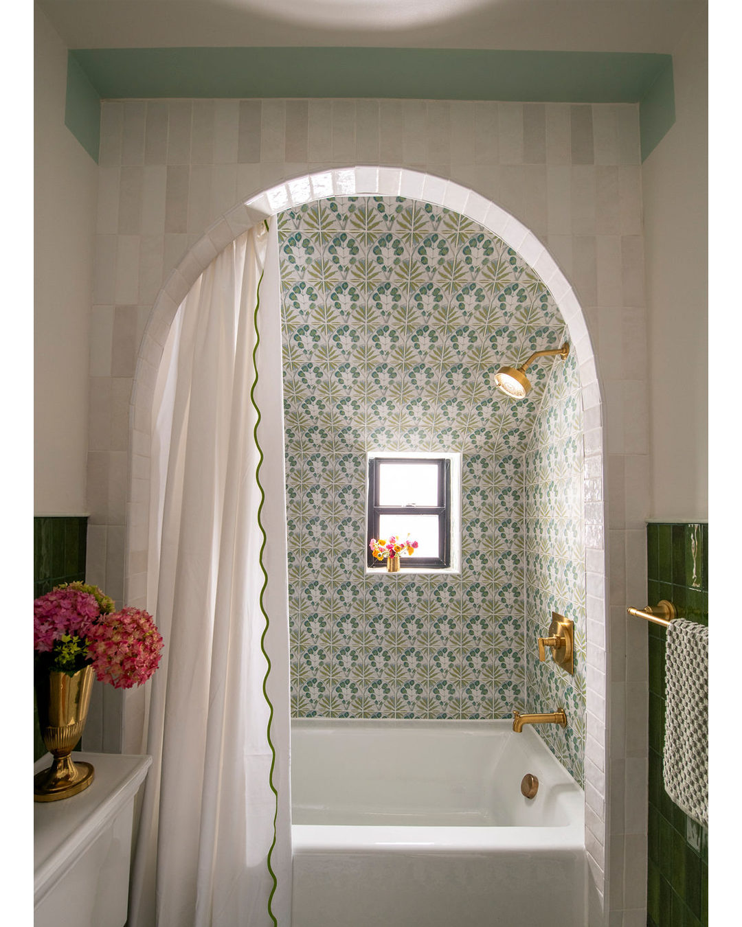 Bathroom with tiled arch shower area, floral patterned tiles, small window with flowers, white bathtub, gold fixtures, showerhead, and curtain with green trim, green and white wall tiles.