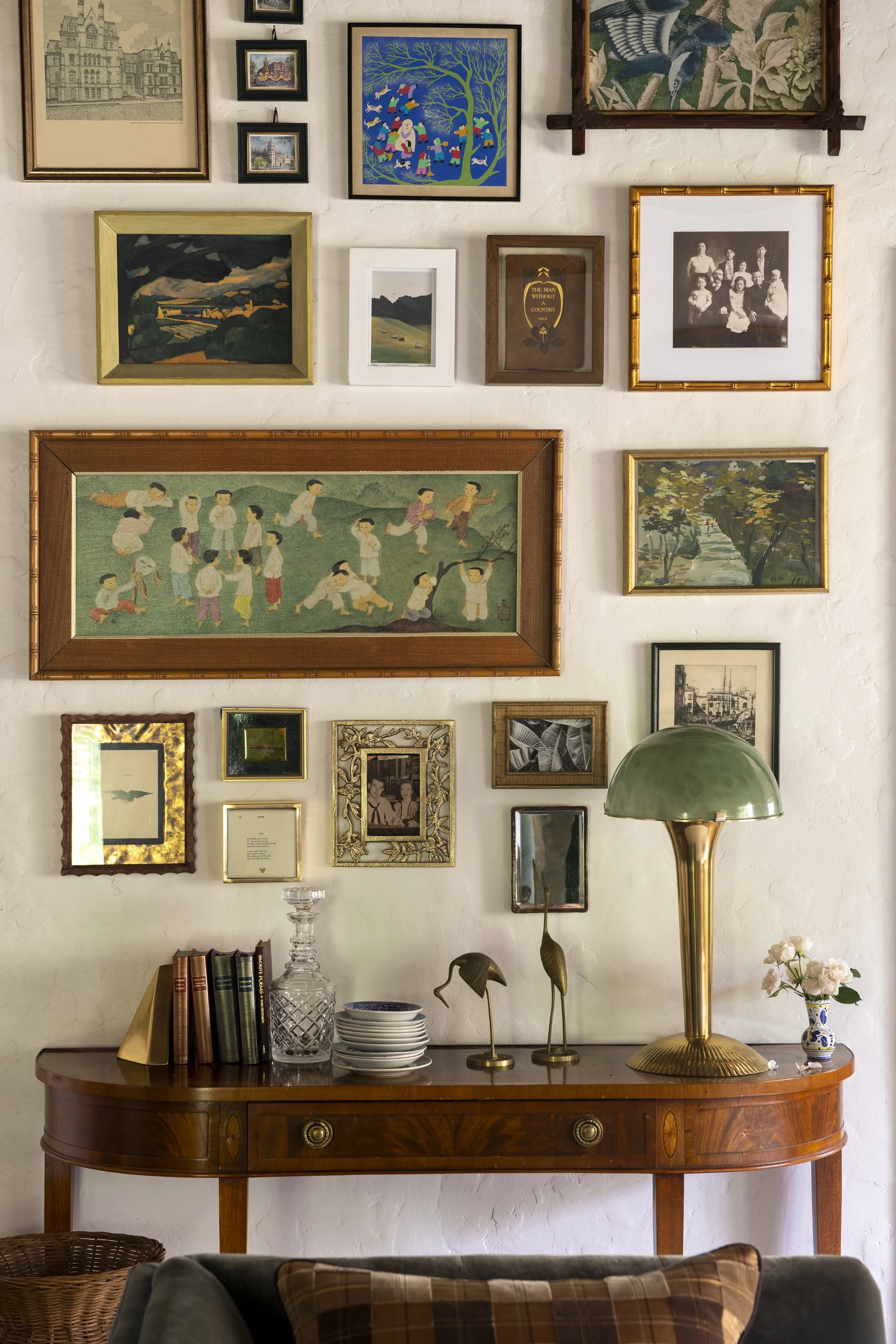 Photo of a wall decorated with various framed pictures and paintings, a wooden sideboard with decorative items, books, and glassware, and a green and gold table lamp.